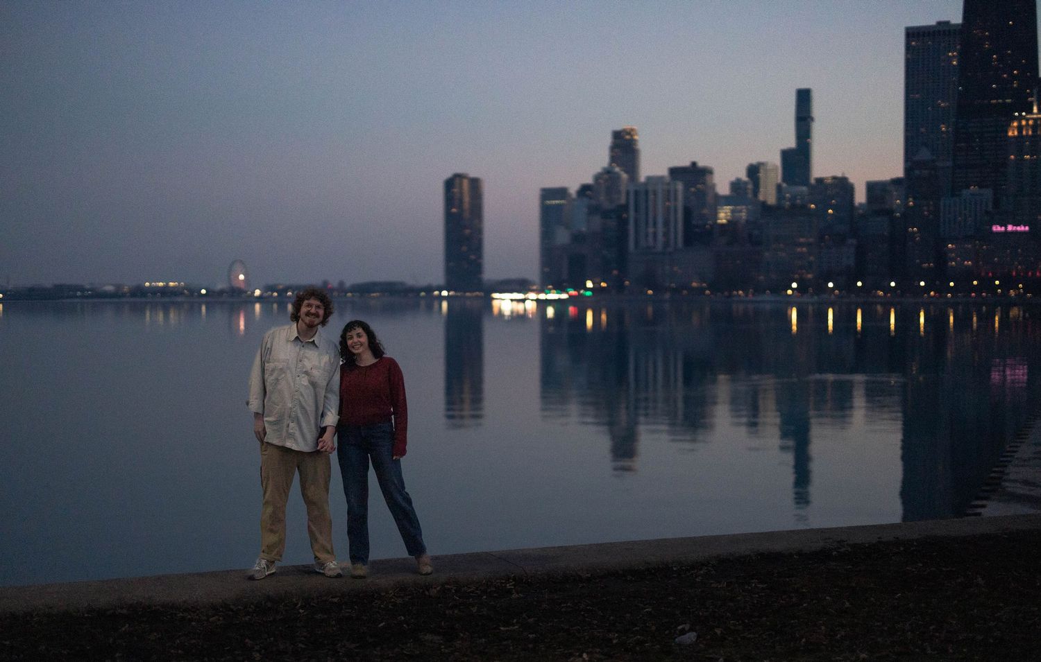Couple standing by lakeside at dusk with Chicago skyline reflecting on calm water in background.