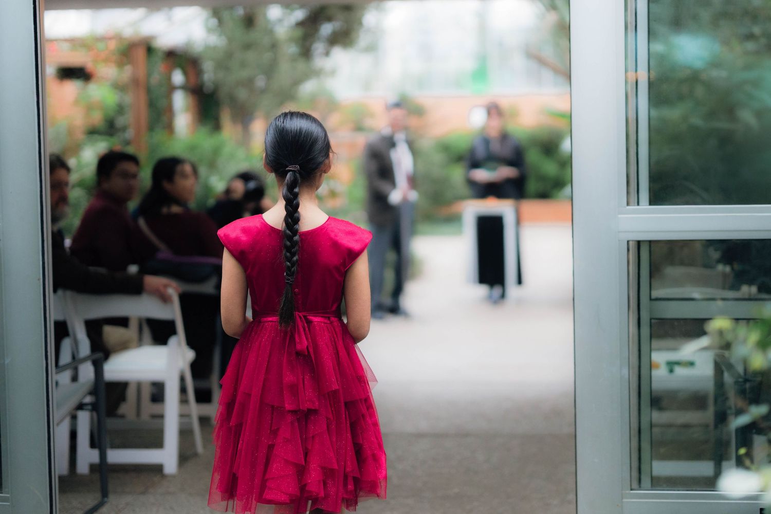Young girl in red dress stands at doorway watching outdoor ceremony with seated guests.