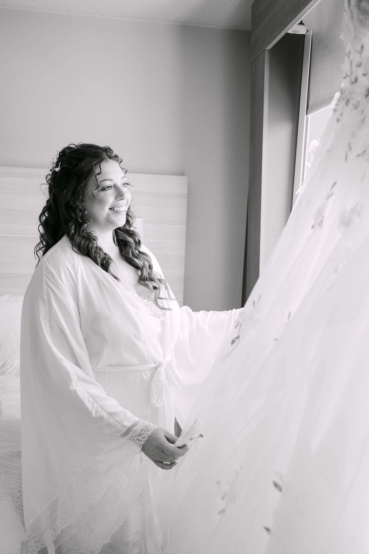Black and white photo of a smiling bride in a white robe admiring her wedding dress hanging by a window.