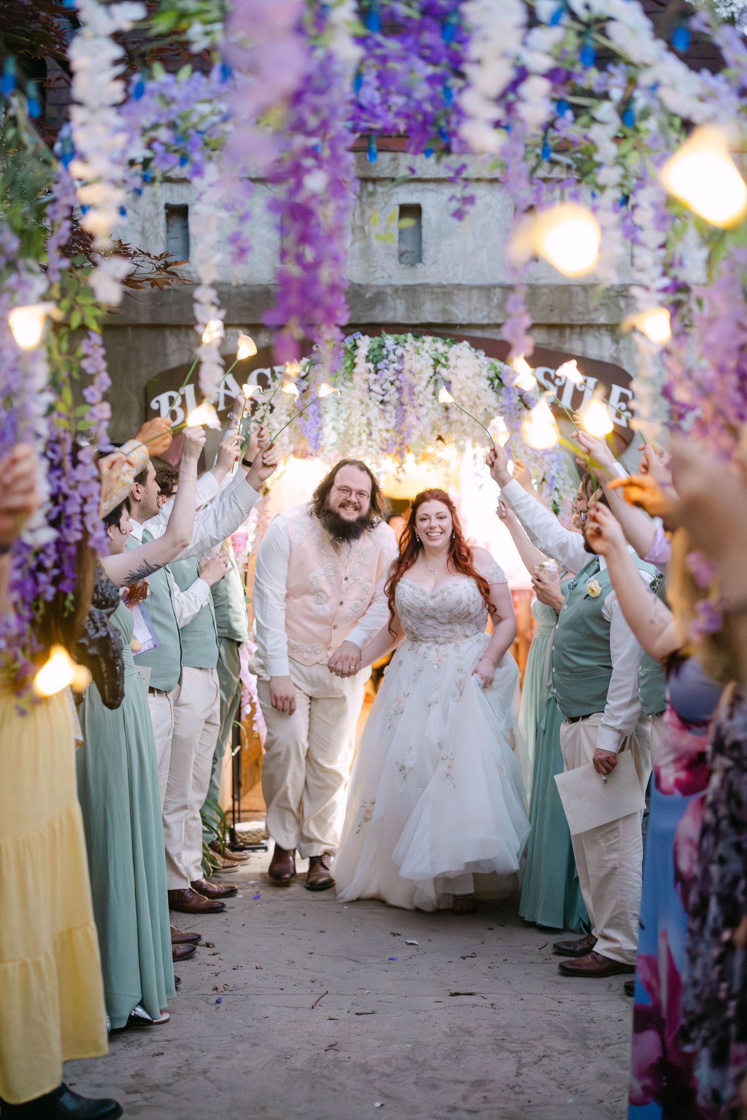 Bride and groom walk through sparkler tunnel under purple wisteria floral arch surrounded by wedding guests.