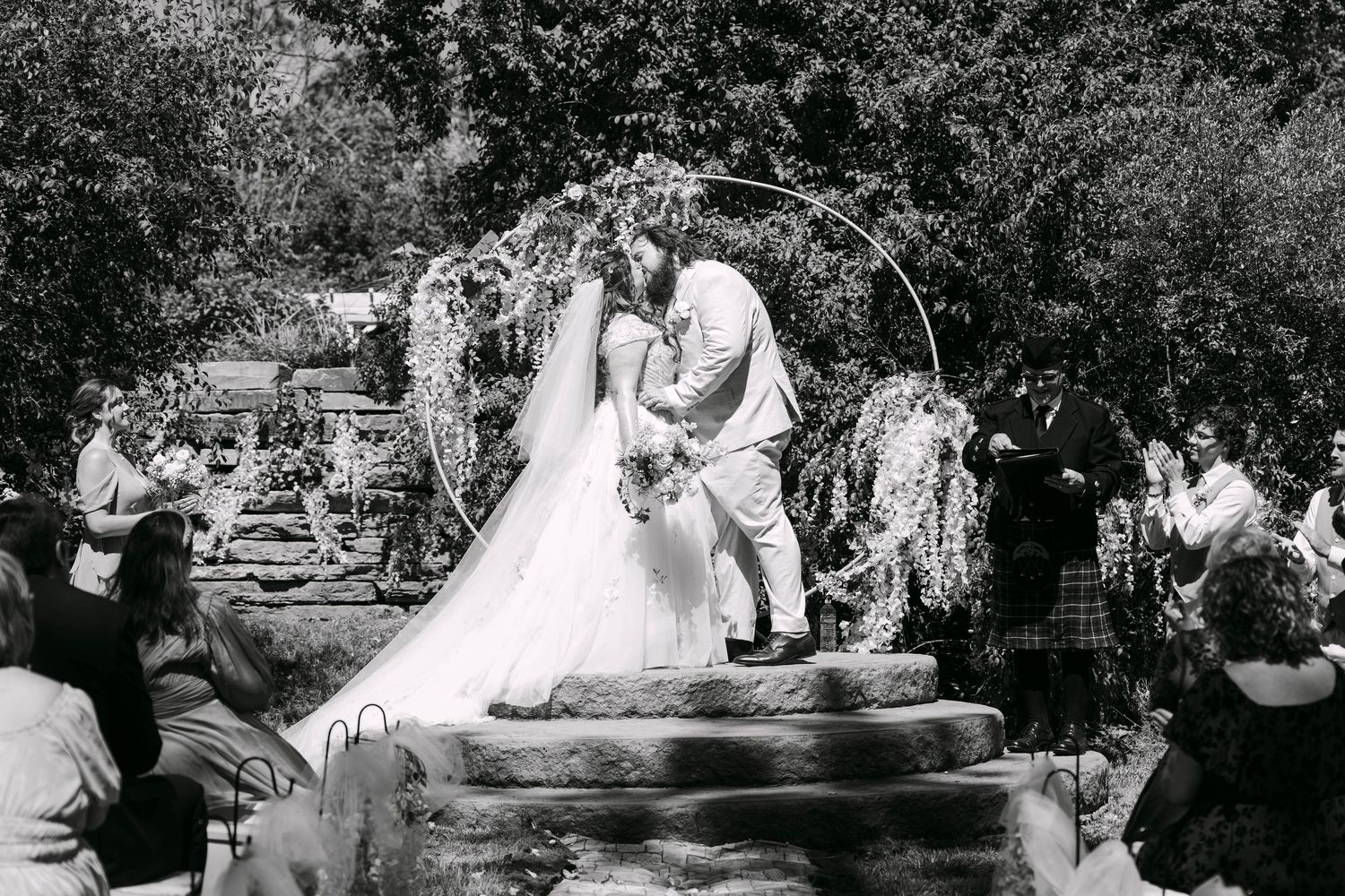 Black and white photo of a couple kissing under a floral arch during an outdoor wedding ceremony with guests watching.
