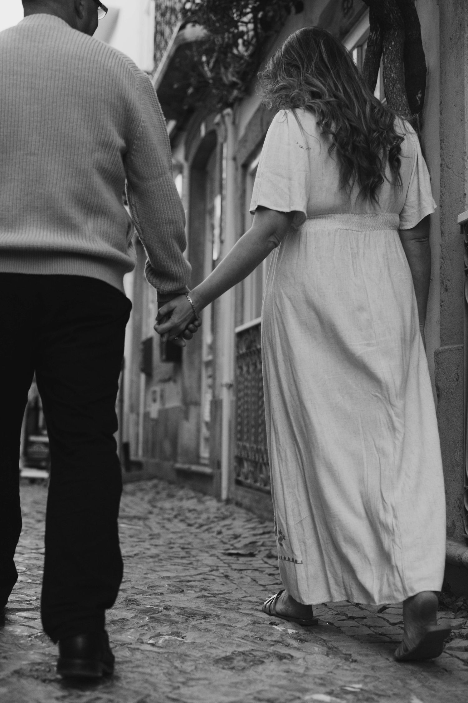 Black and white photo of a couple holding hands while walking down a cobblestone street, woman in white dress.