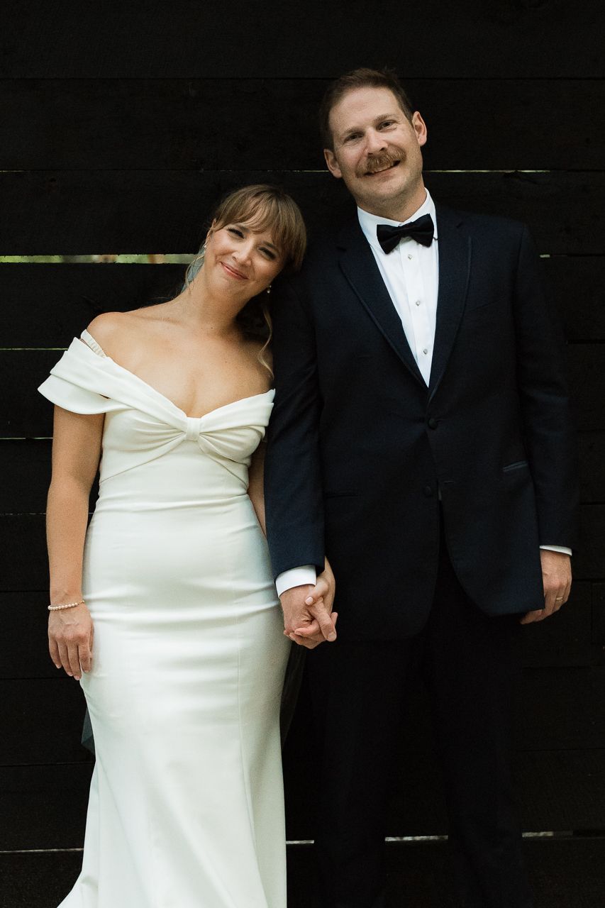 Elegant couple in formal black and white wedding attire pose against dark background.
