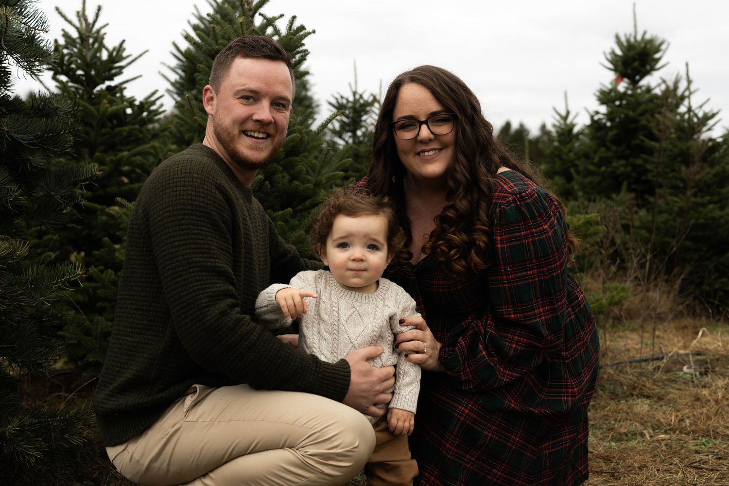 Family of three posing among Christmas trees at a tree farm, parents smiling with baby in festive holiday attire.