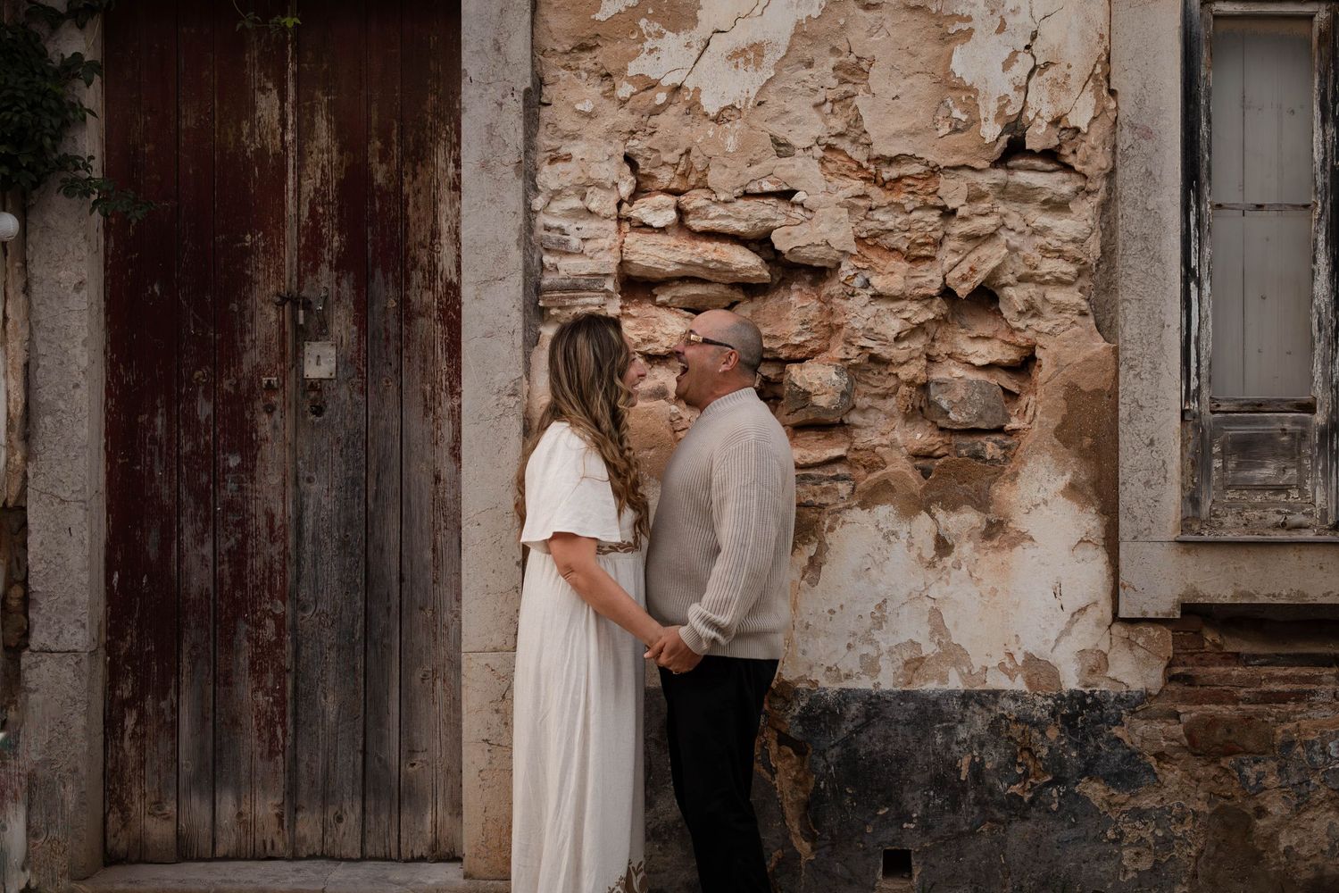 Couple holding hands and facing each other in front of a rustic stone wall and weathered wooden door.
