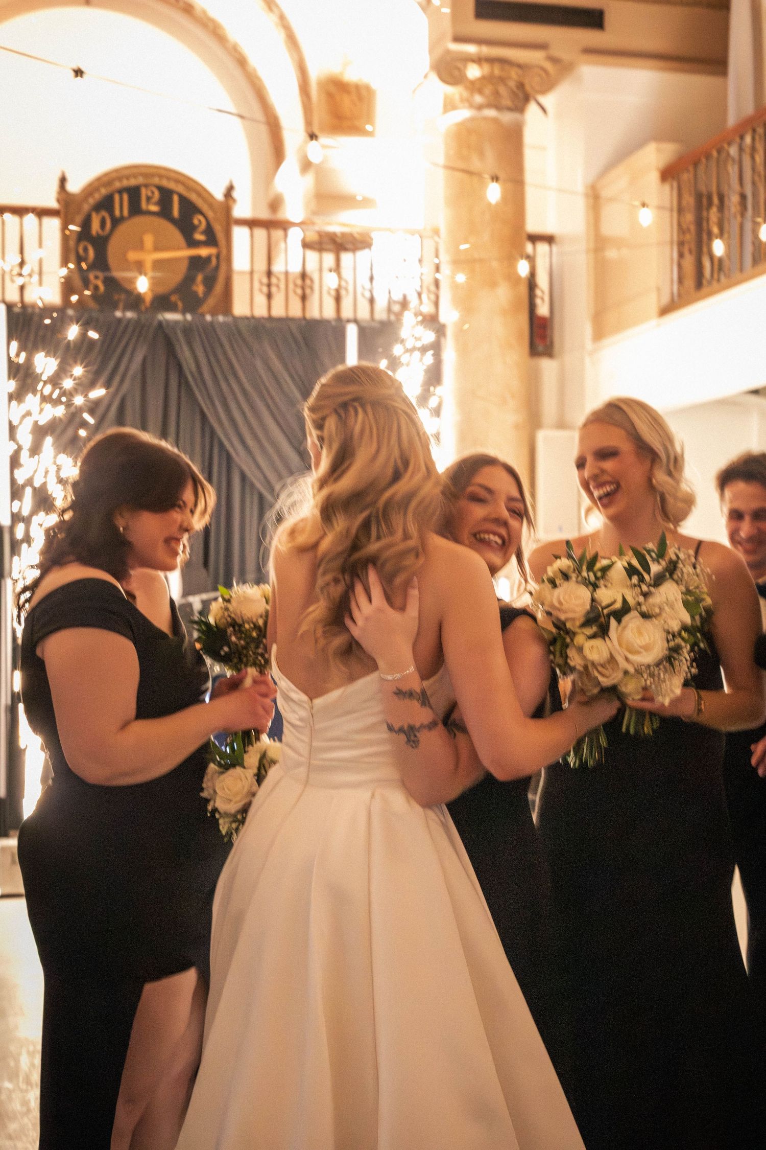 Bride in white gown laughing with bridesmaids in black dresses holding white bouquets at elegant wedding reception.
