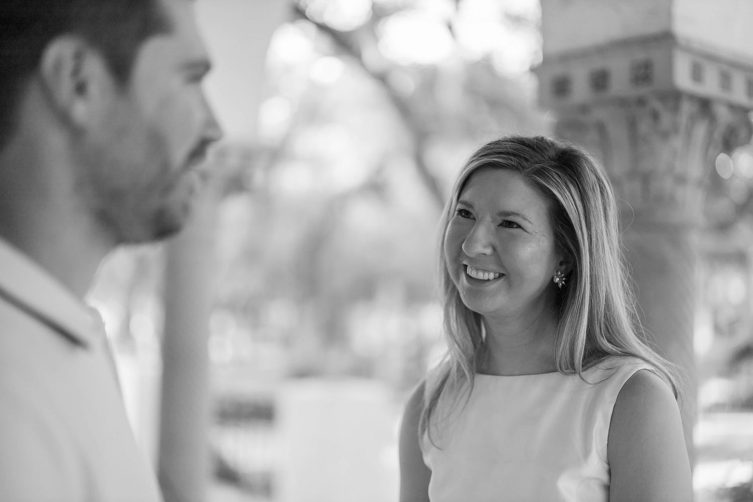 Black and white photo of a smiling woman in a sleeveless top conversing with a man outdoors.
