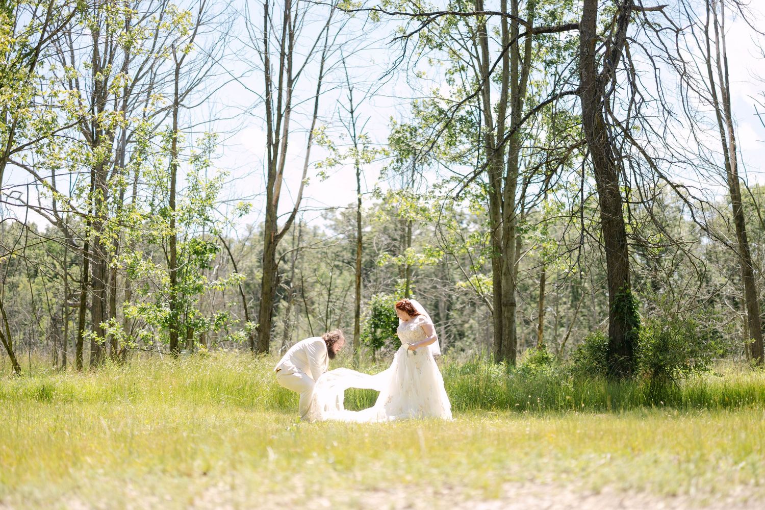 Bride in white gown with bridesmaid in sunny forest clearing, surrounded by tall trees and green grass.