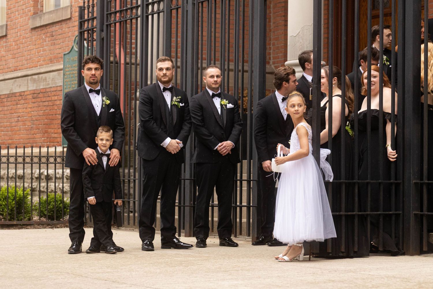 Wedding party with groomsmen in tuxedos and a flower girl in white dress standing outside ornate iron gates.