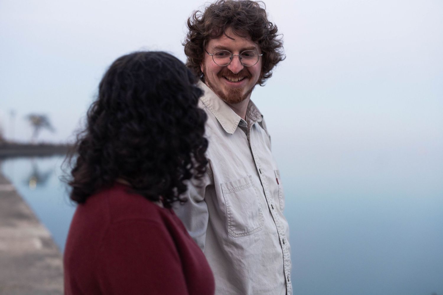 A smiling curly-haired man in a white shirt stands near waterfront, facing a woman with dark curly hair in a red top.
