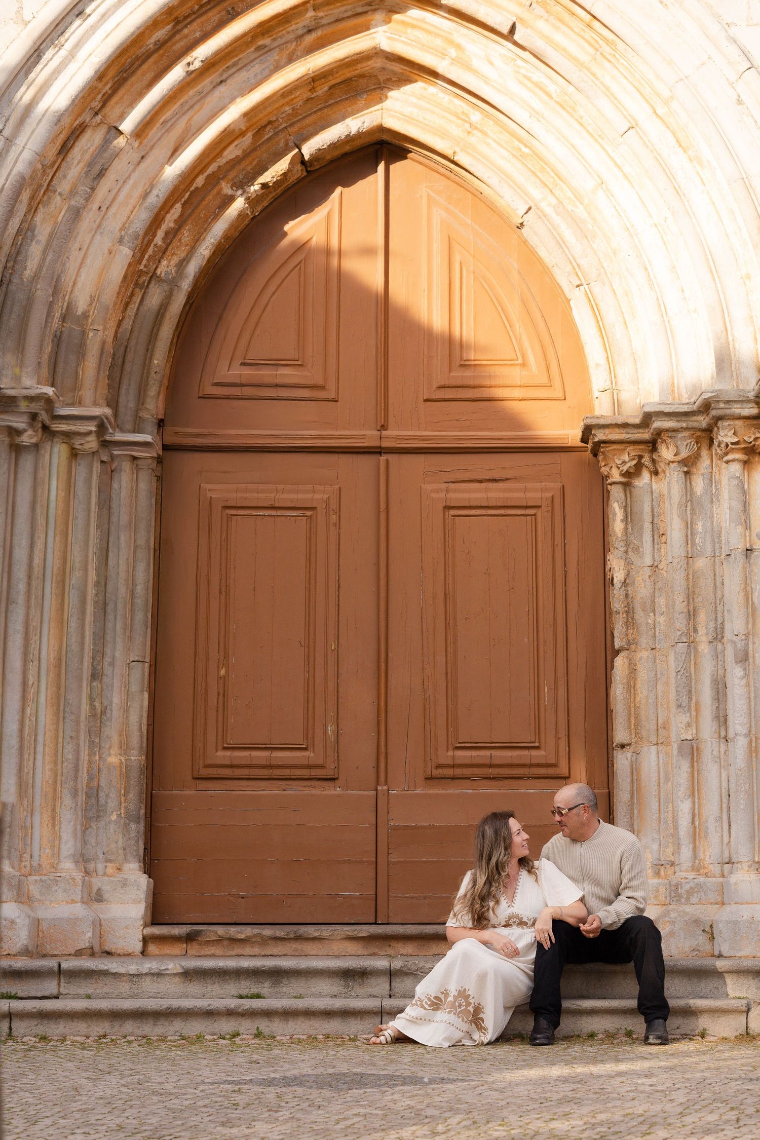 Couple sitting on steps in front of a large wooden arched church door, sharing a tender moment.
