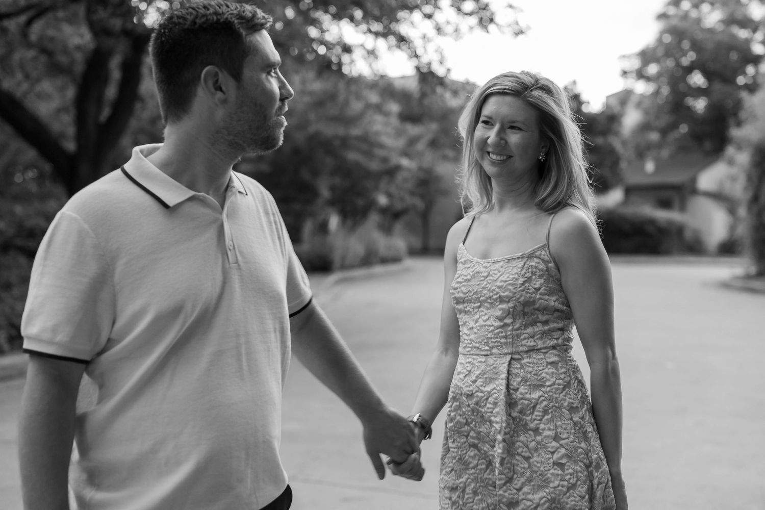 Black and white photo of a smiling couple holding hands on a tree-lined street.