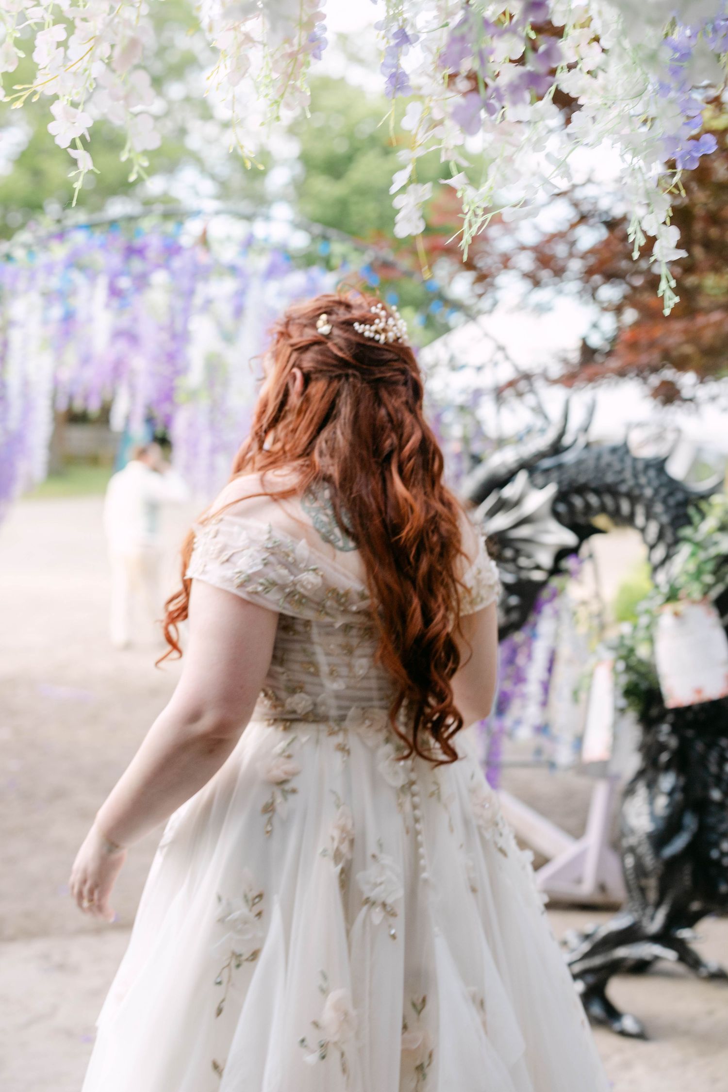 Red-haired bride in floral wedding gown stands under wisteria arch at outdoor fantasy wedding ceremony.