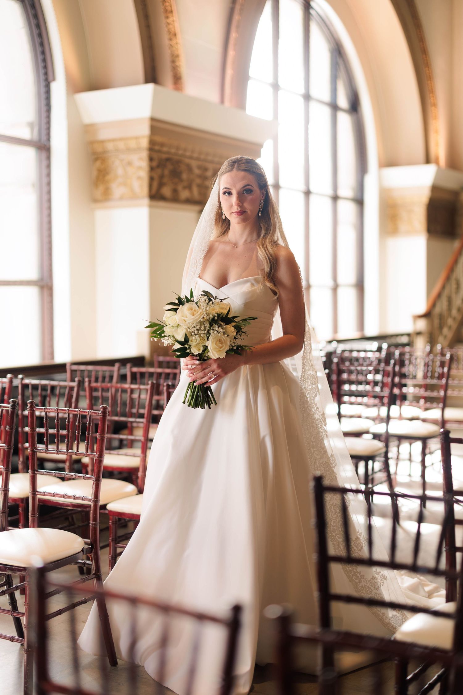 Elegant bride in strapless gown holds white bouquet beside arched windows in a grand venue.