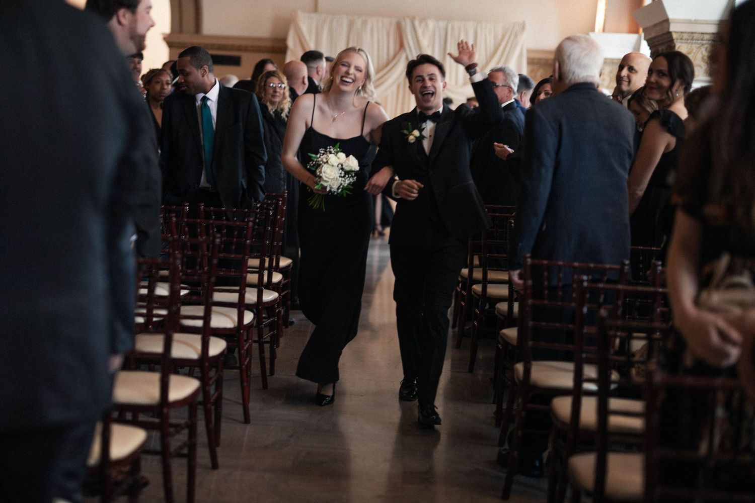 Joyful couple walks down wedding aisle in elegant venue, bride in white gown, escort in black suit, guests celebrating.
