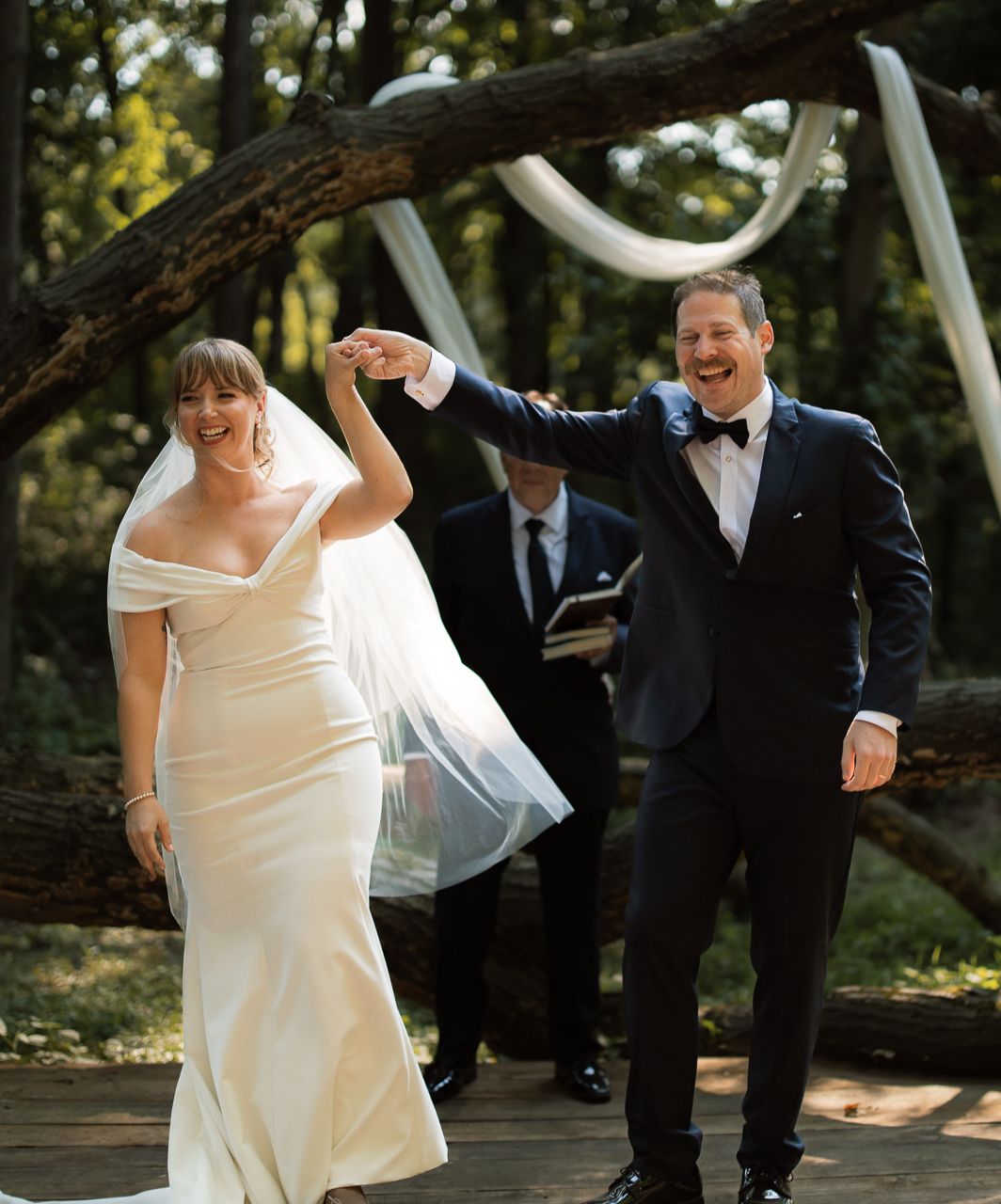 Joyful newlyweds walk back down aisle after outdoor forest wedding ceremony, groom spinning bride's hand.