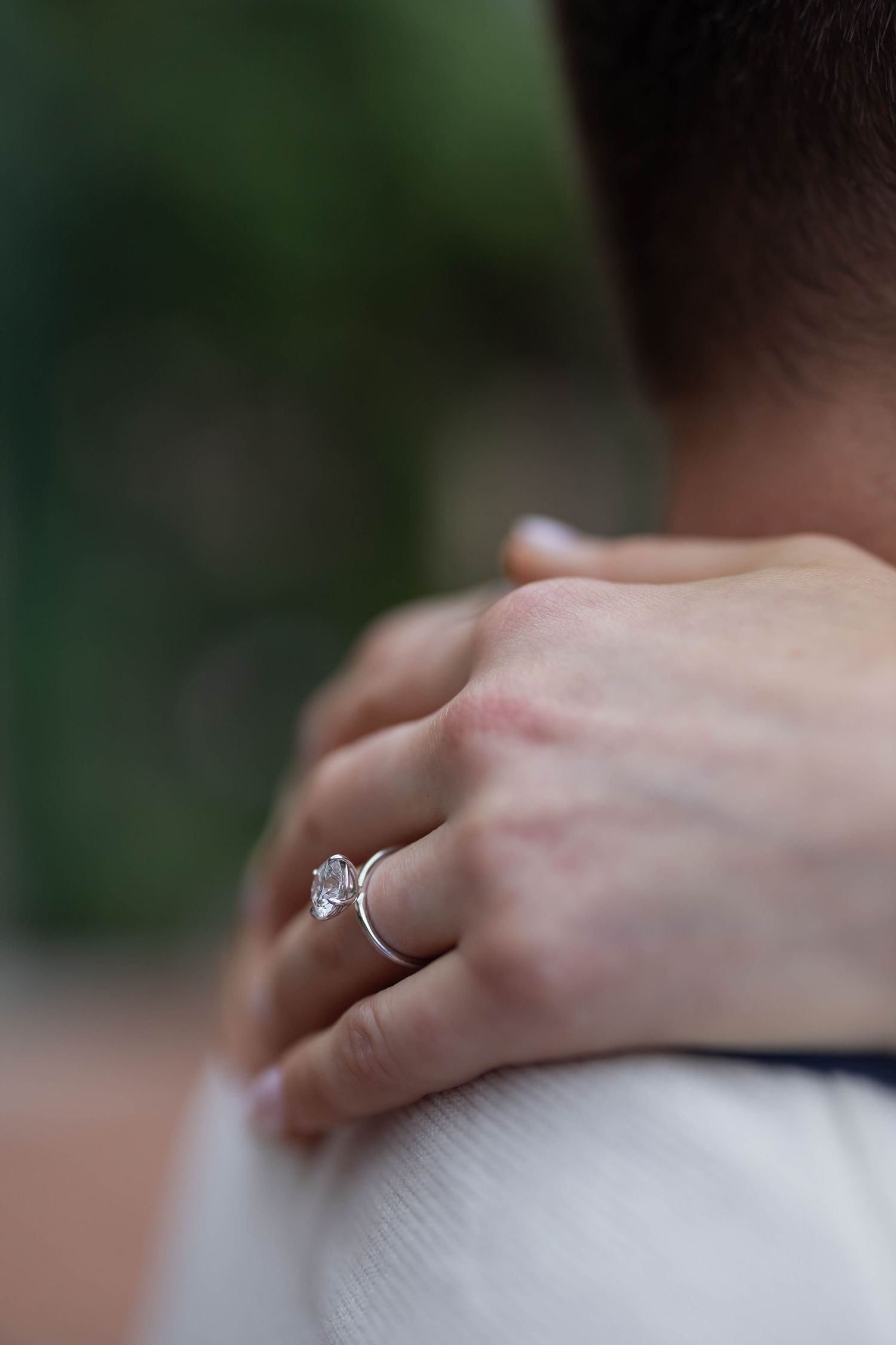 A close-up of a diamond engagement ring on a woman's finger as she rests her hand on a man's shoulder.