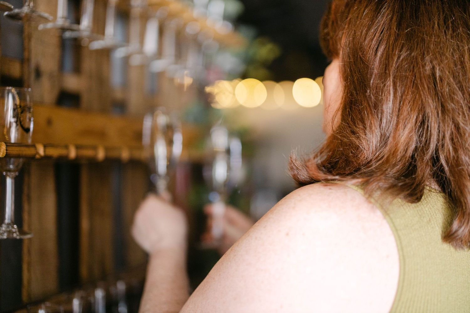 A woman with red hair browsing wine glasses at a rustic bar with warm bokeh lights in the background.