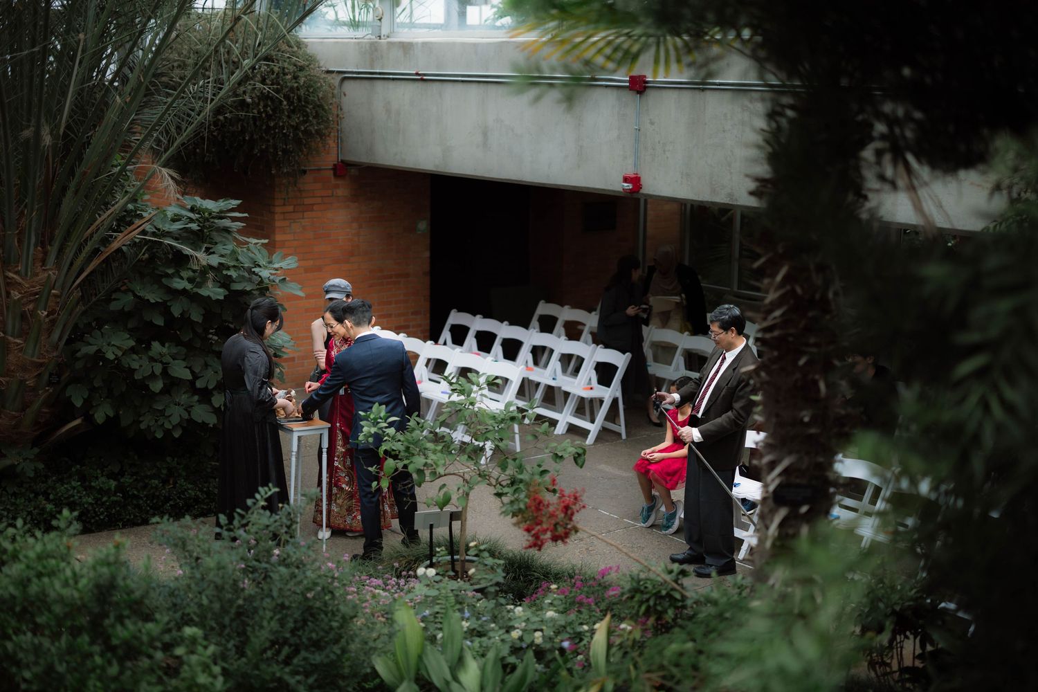 Guests gather near white chairs set up for an outdoor wedding ceremony surrounded by lush greenery.