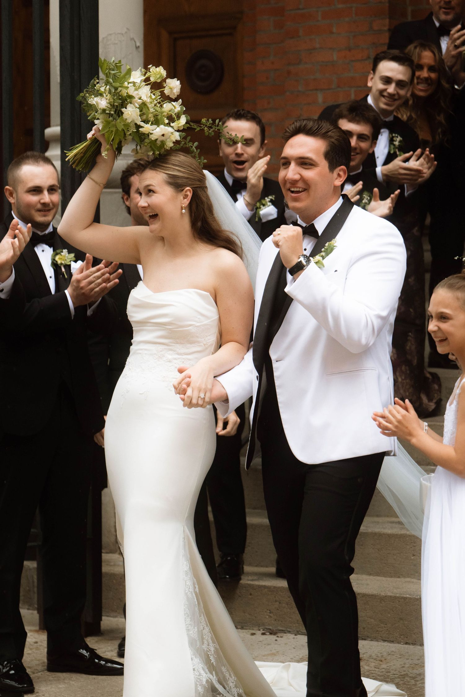 Joyful bride and groom exit church steps celebrating with wedding party in black and white tuxedos.