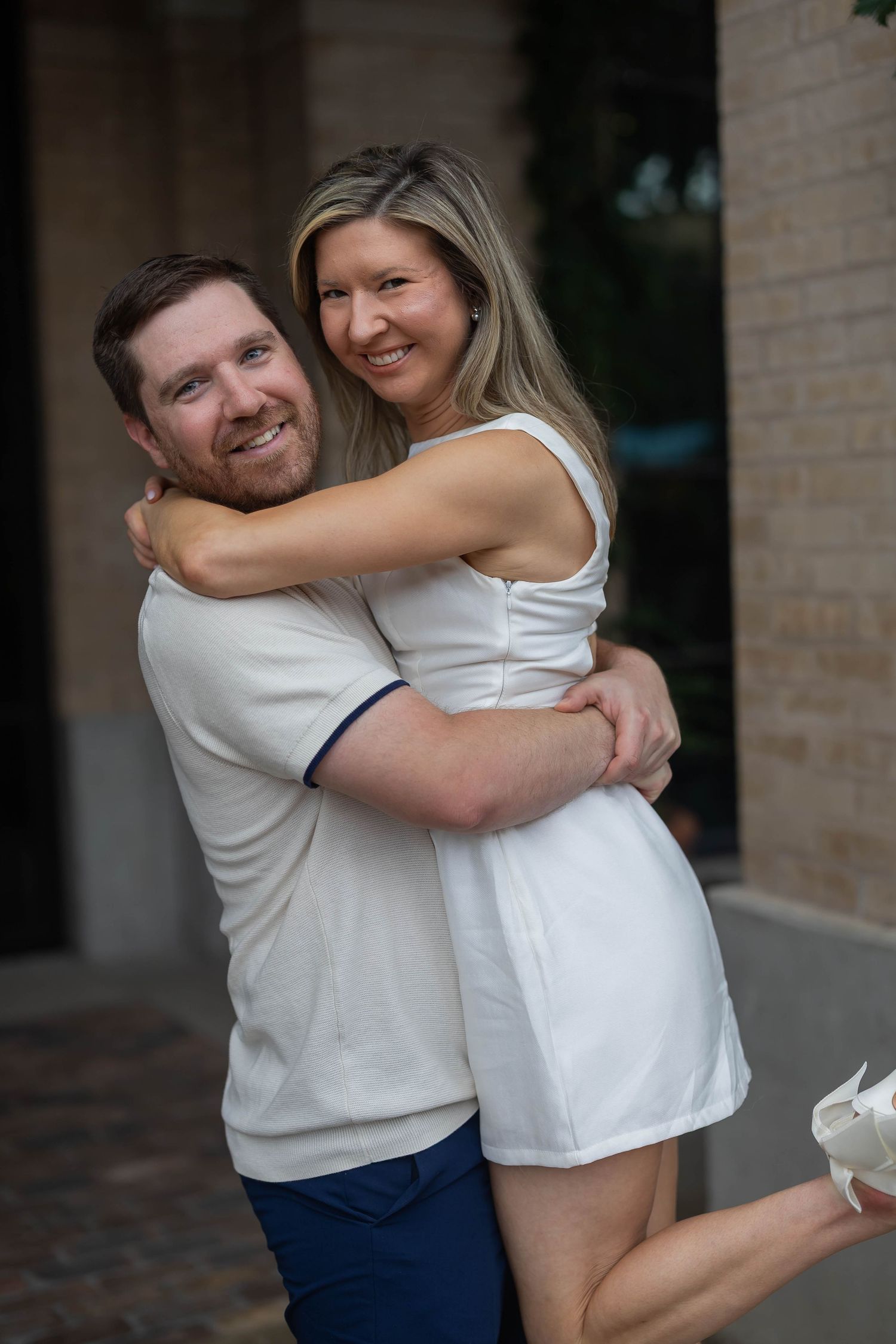 Happy couple embracing outdoors, woman in white dress hugging smiling man in white polo and blue shorts.