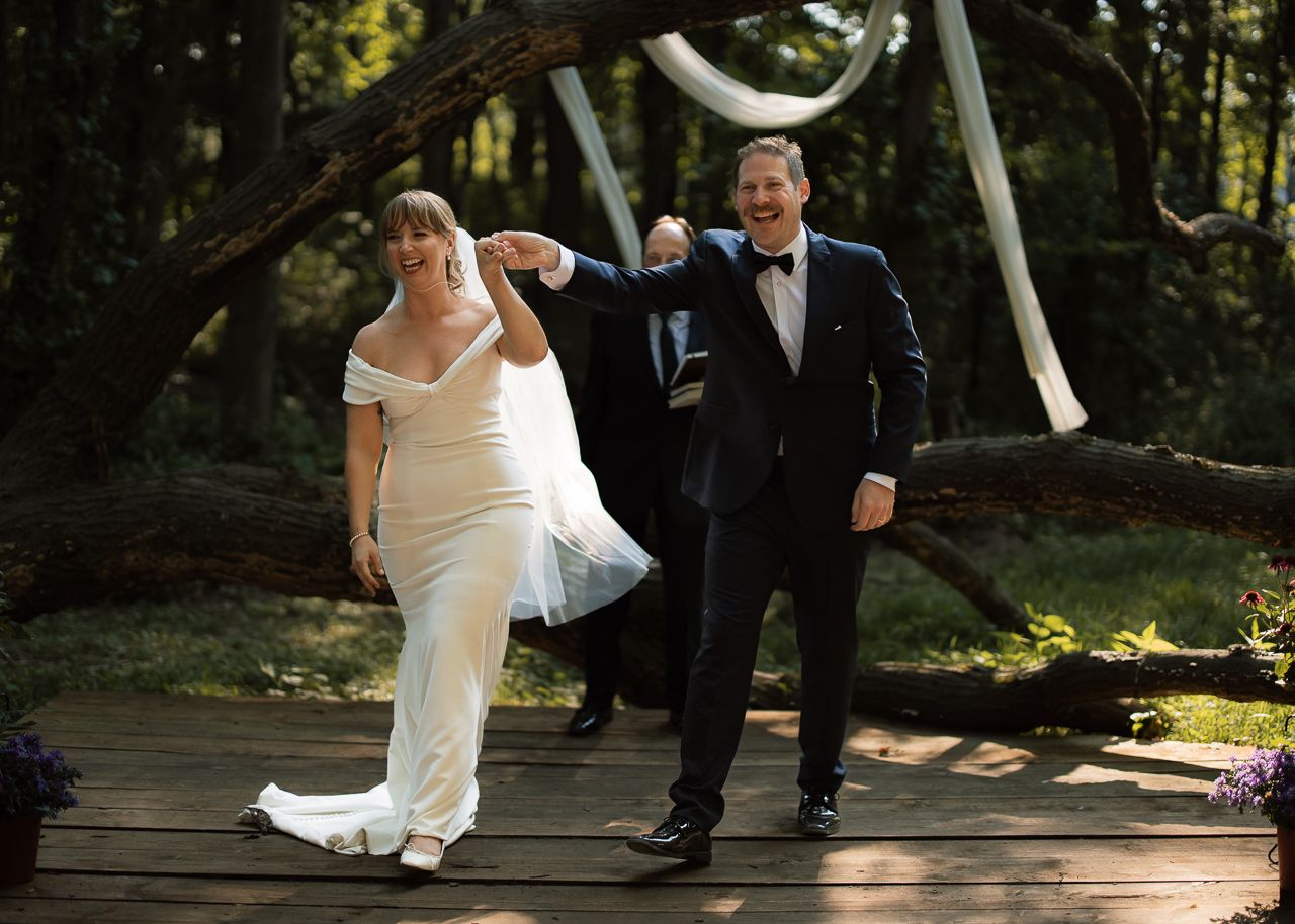 Bride and groom walking happily on wooden deck at outdoor forest wedding ceremony with arch backdrop.