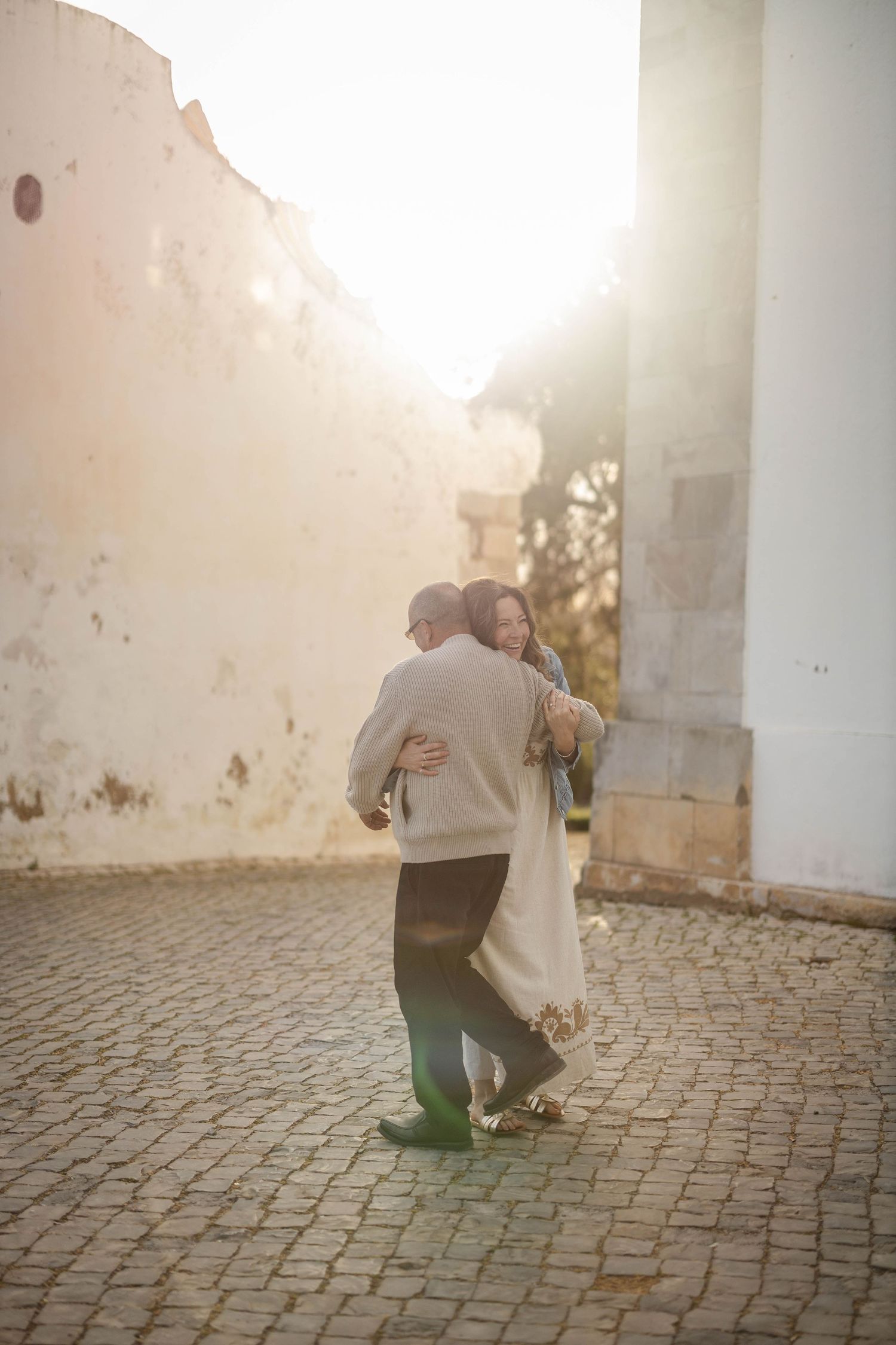 Couple embracing and dancing on cobblestone street bathed in warm golden sunlight near rustic white walls.