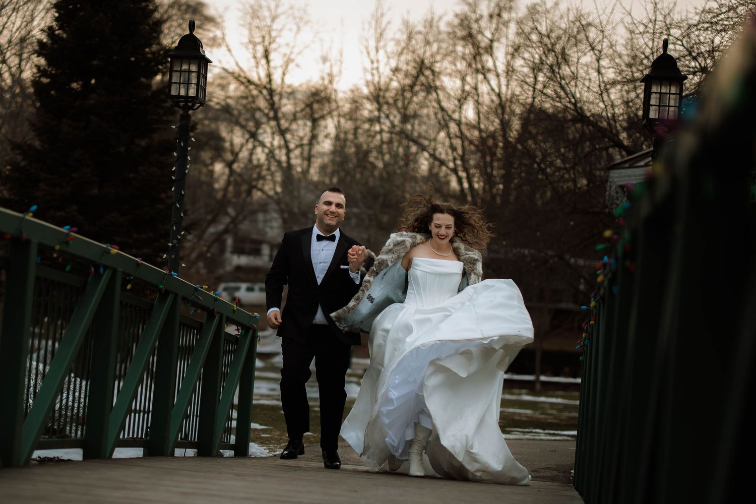 Bride and groom joyfully running on a bridge in winter, bride lifting her white gown, groom in black tuxedo.