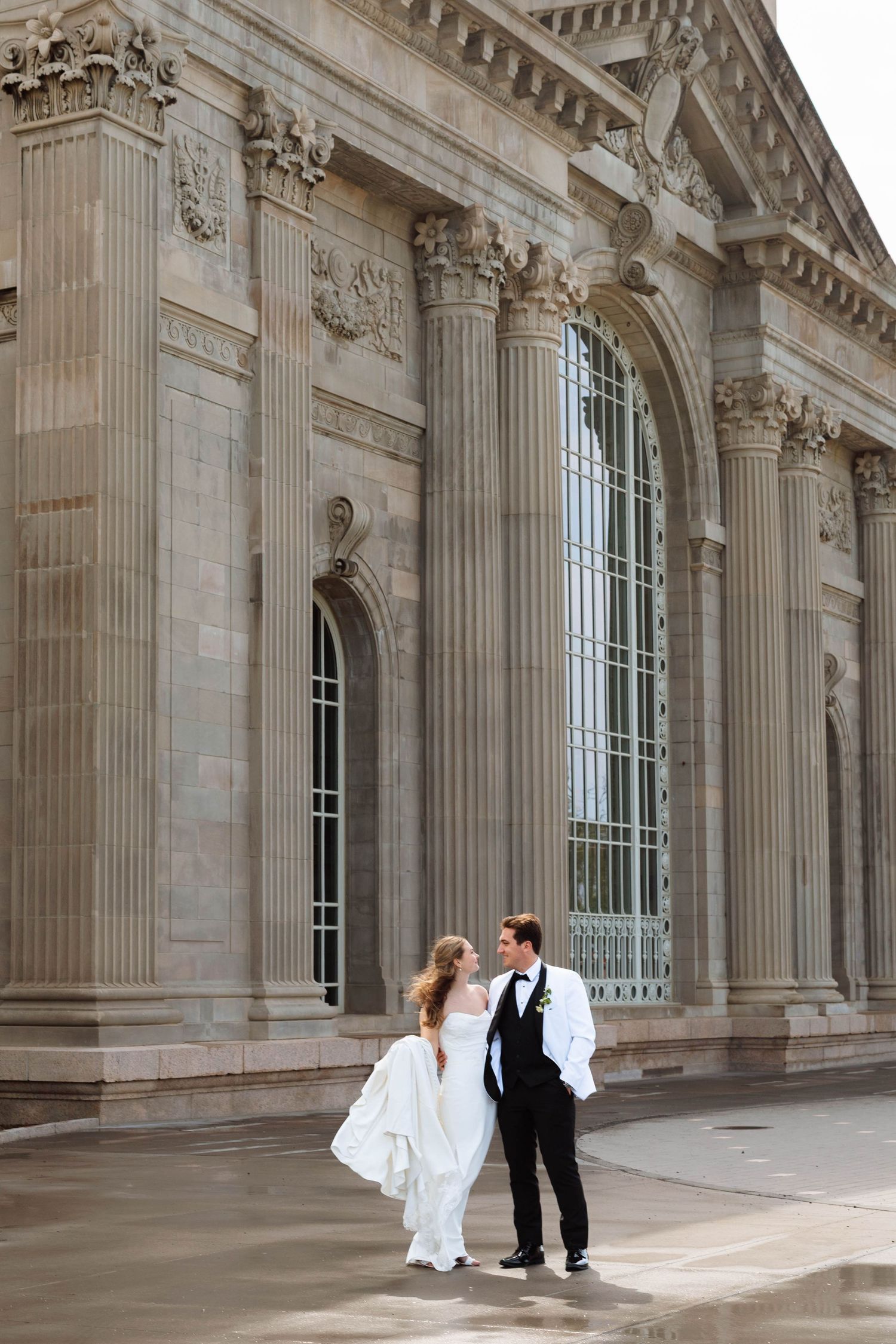 Bride and groom walking hand in hand in front of a grand neoclassical building with tall columns.