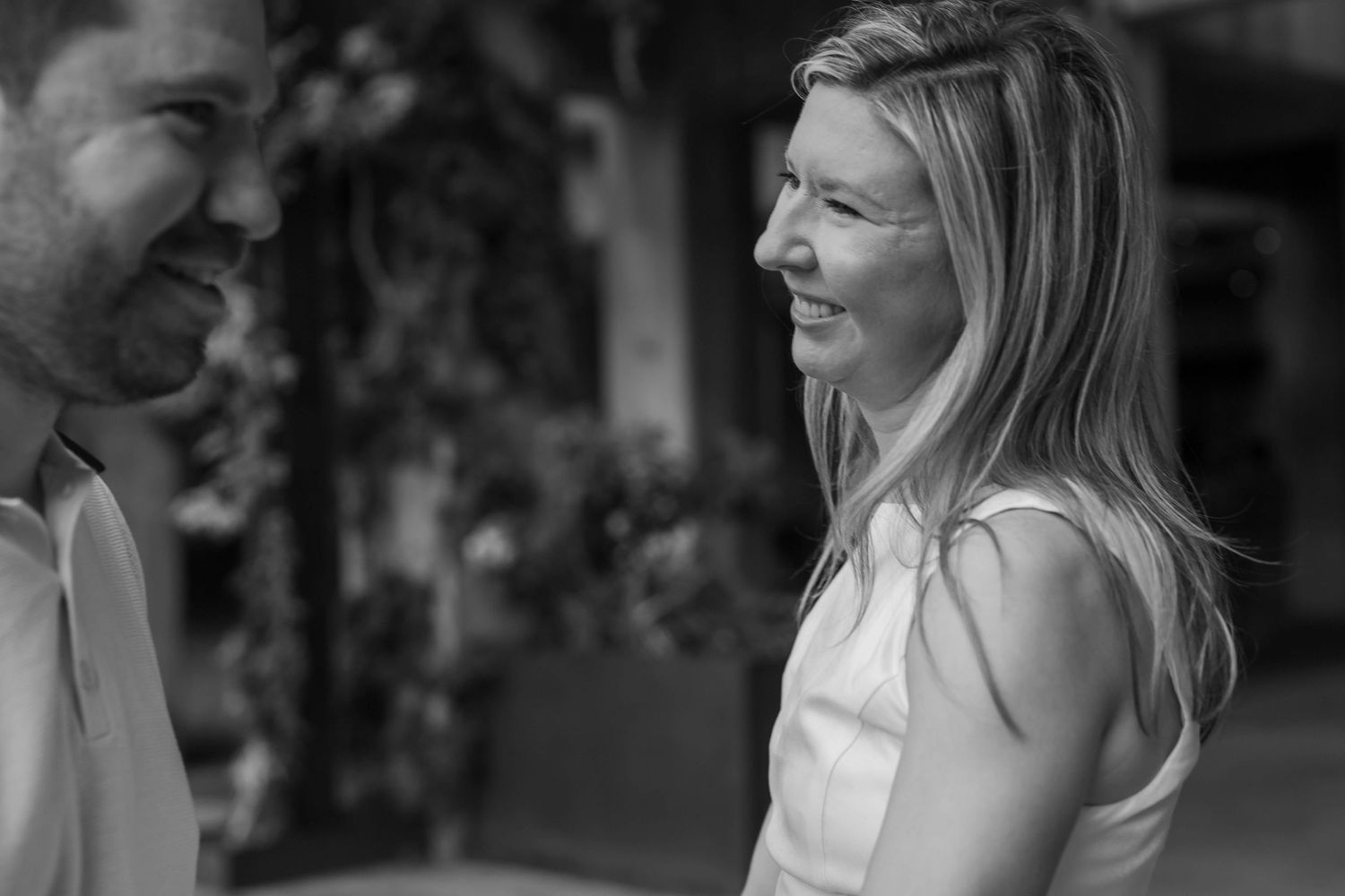 Black and white photo of a smiling couple sharing a joyful moment outdoors, with blurred greenery in the background.