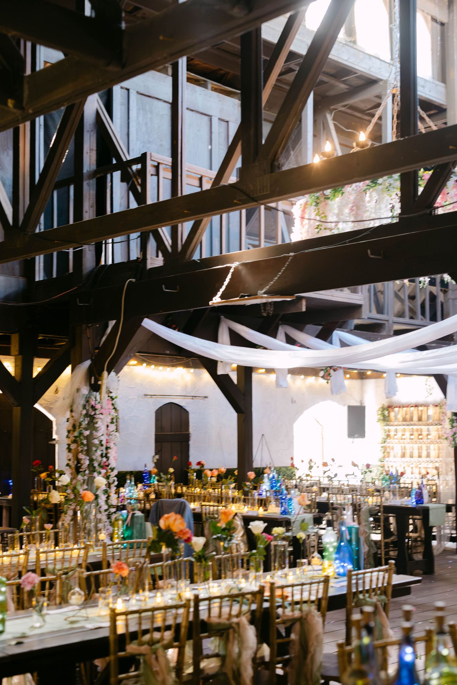 Elegant wedding reception in industrial venue with string lights, draped fabric, colorful floral centerpieces, and gold chiavari chairs.