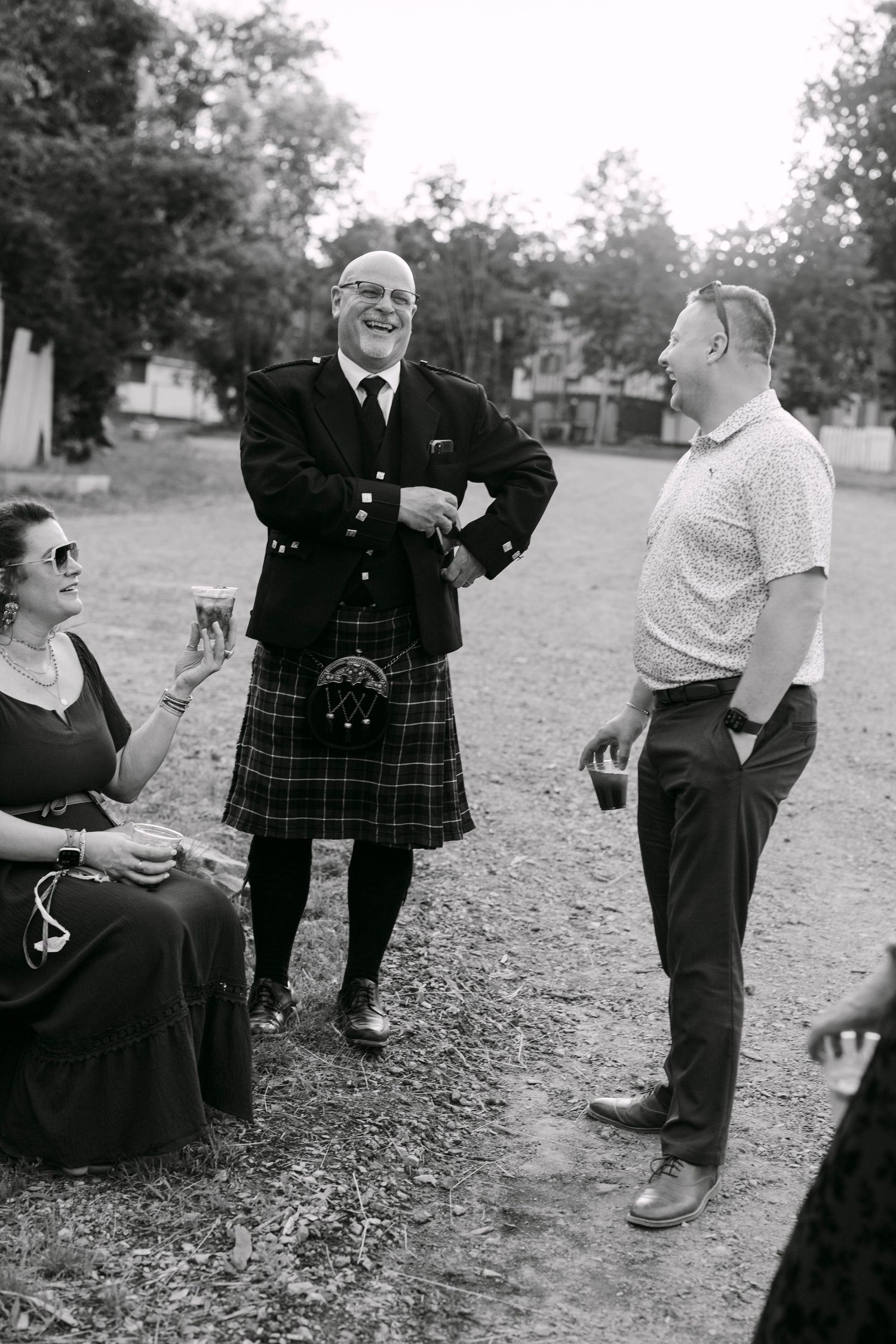 Black and white photo of a man in a Scottish kilt laughing with guests at an outdoor wedding reception.