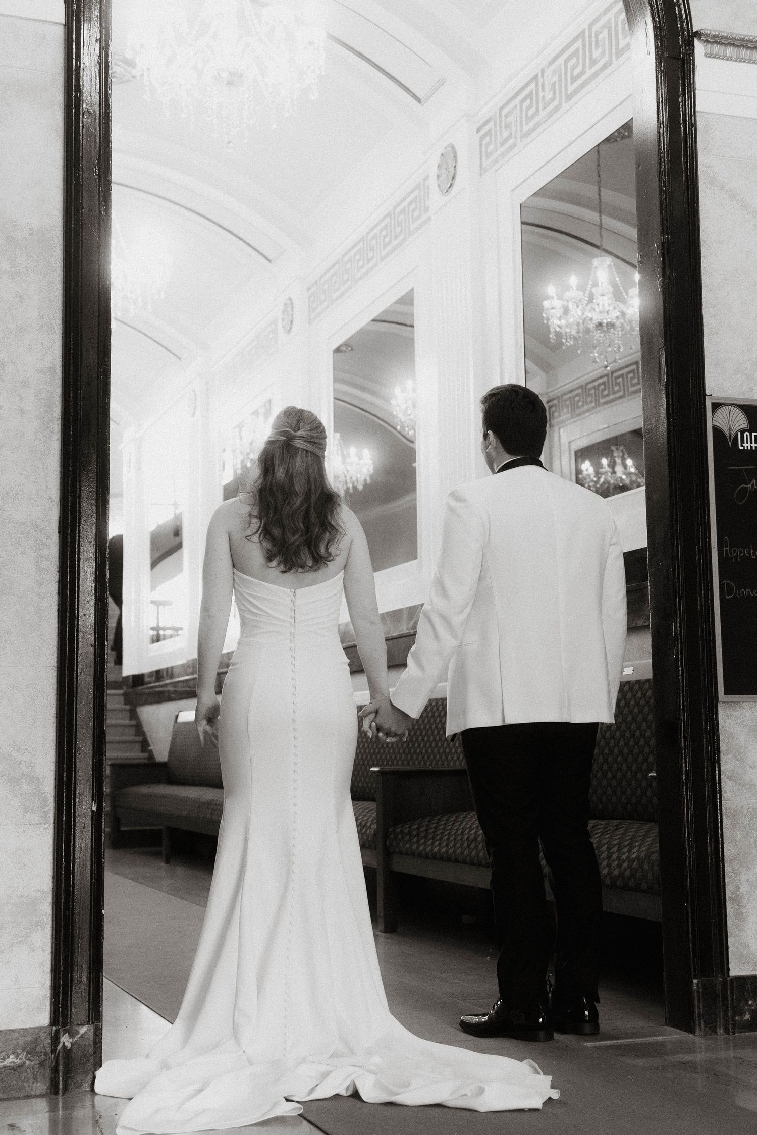 Black and white photo of bride and groom holding hands at venue entrance.