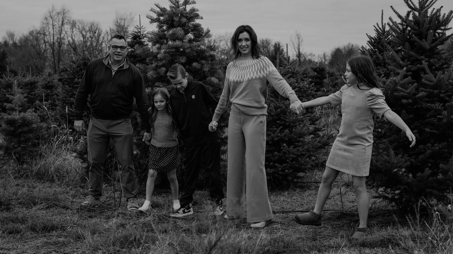 Black and white family photo at a Christmas tree farm with parents and three children holding hands outdoors.