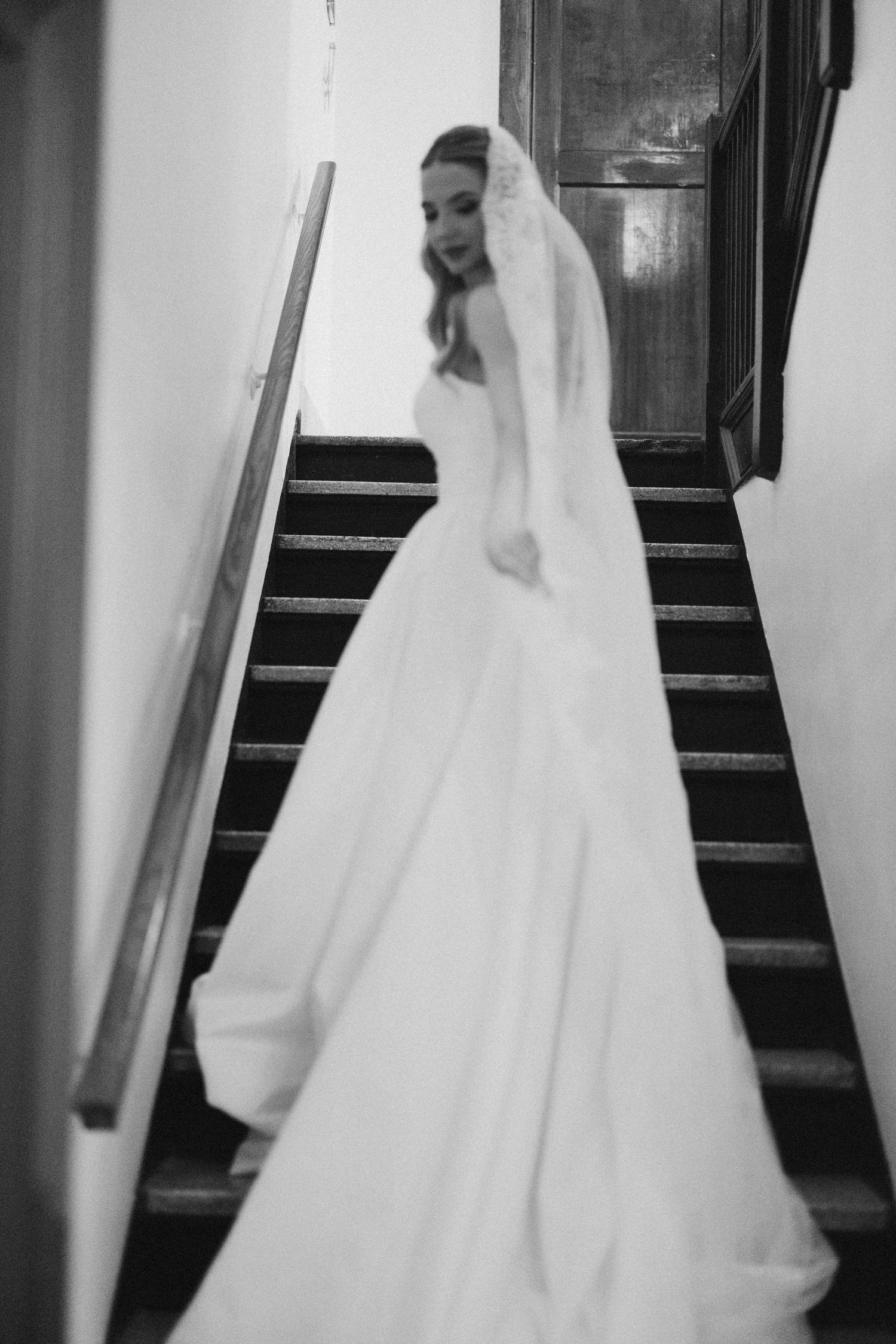 Black and white photo of bride in white gown and veil descending wooden staircase in elegant venue.