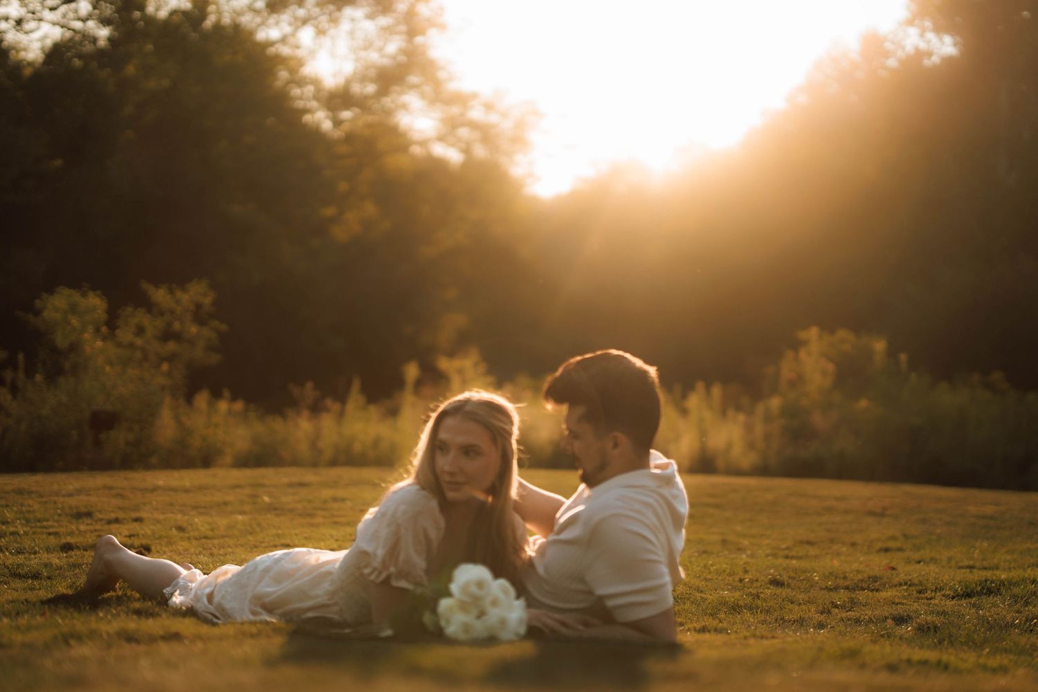 A couple lays in a sunlit meadow during golden hour as warm sunlight filters through trees in the background.