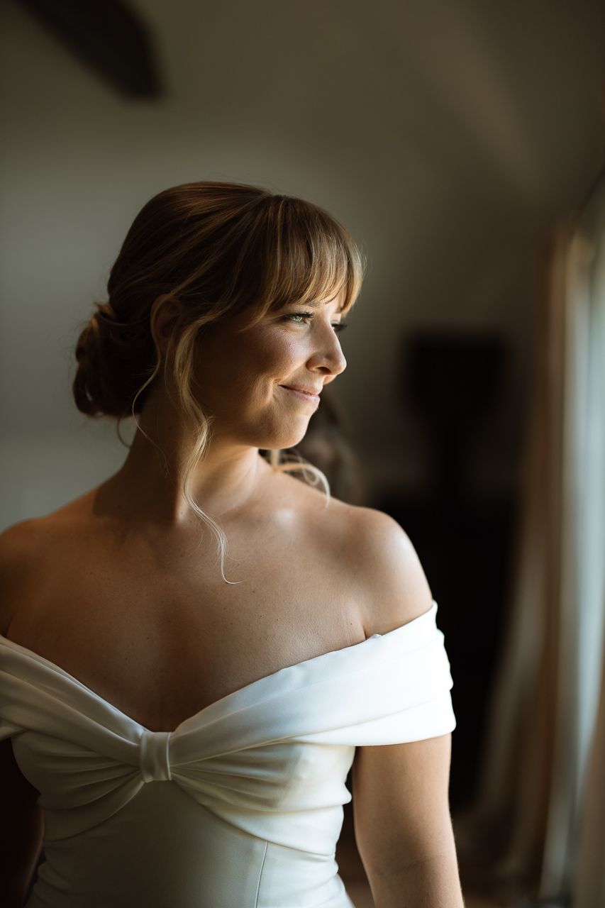 Smiling bride in off-shoulder white gown with updo hairstyle gazing toward window in soft natural light.