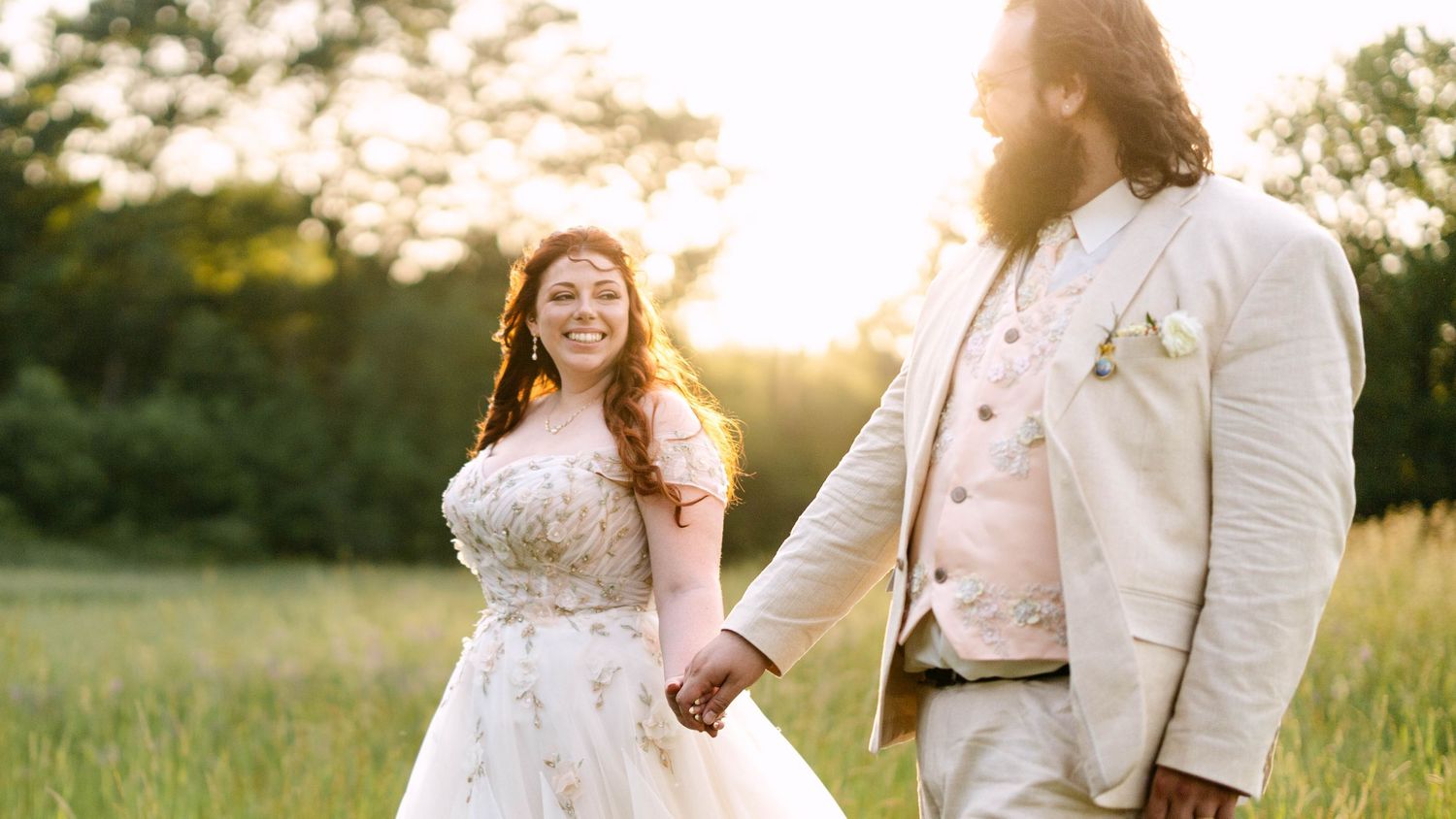 A bride and groom hold hands in a sunlit meadow, she in a floral gown, he in a beige suit with a long beard.
