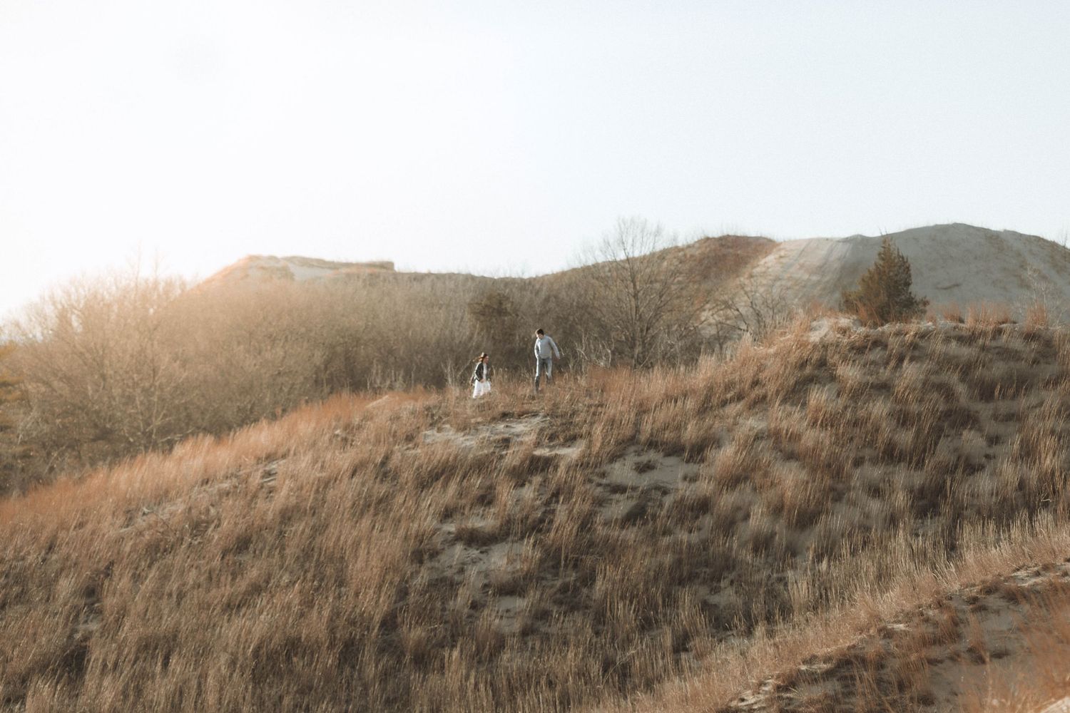 Two hikers walk along a dry golden hillside with sparse vegetation under a bright hazy sky.