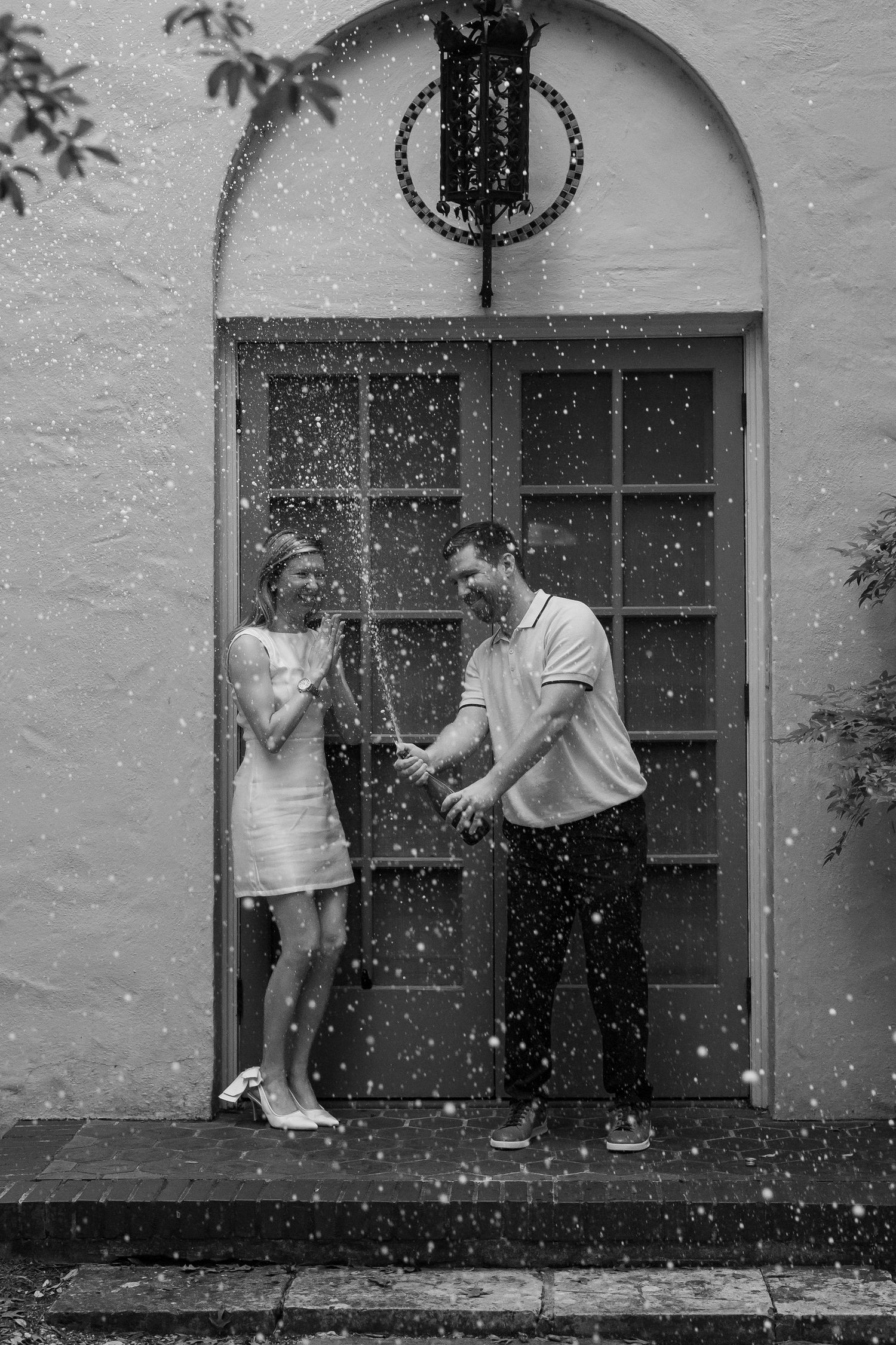 Black and white photo of couple popping champagne bottle outside arched doorway.