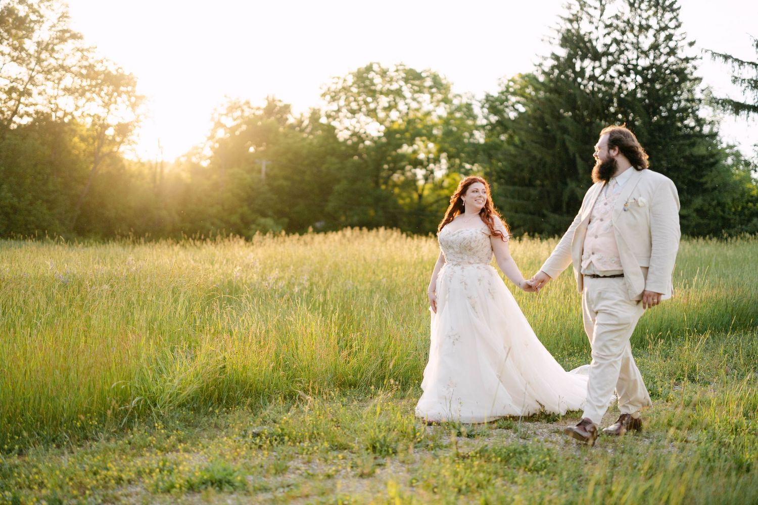 Bride and groom holding hands walking through a sunlit meadow at golden hour in wedding attire.