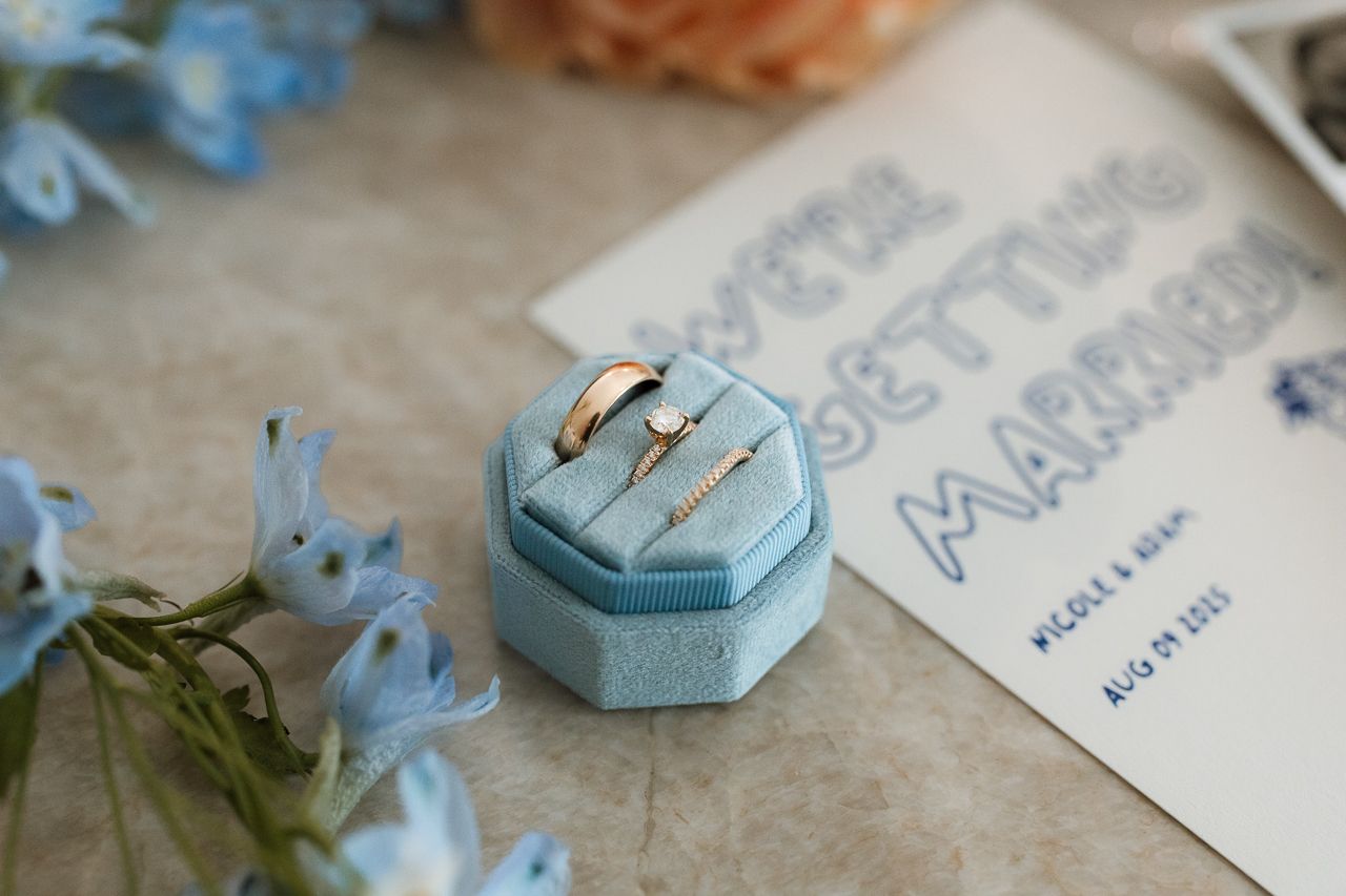 Gold wedding rings displayed in a blue velvet ring box beside a wedding announcement card and delicate blue flowers.