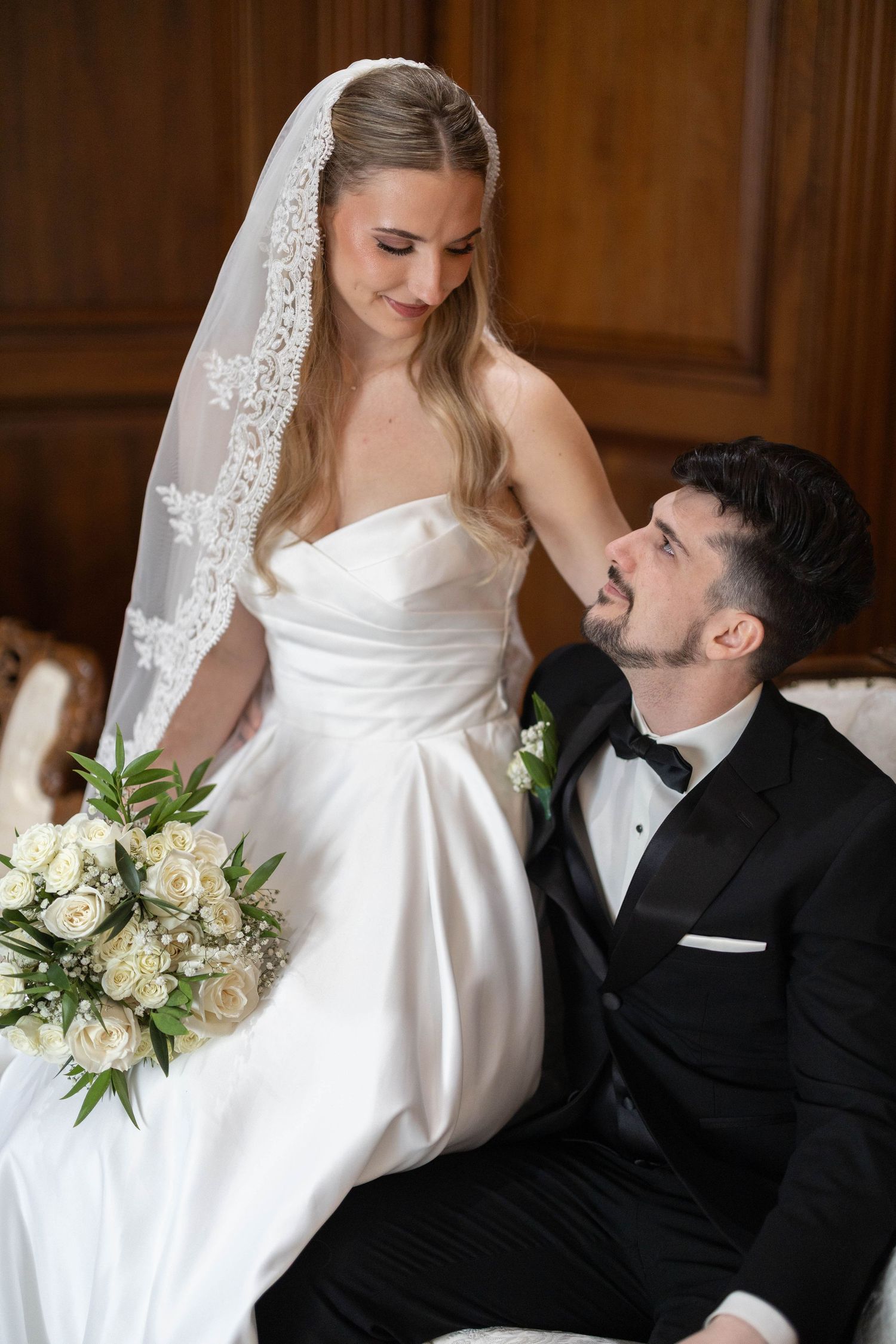 Bride in white strapless gown and lace veil sits with groom in black tuxedo on elegant sofa holding bouquet.
