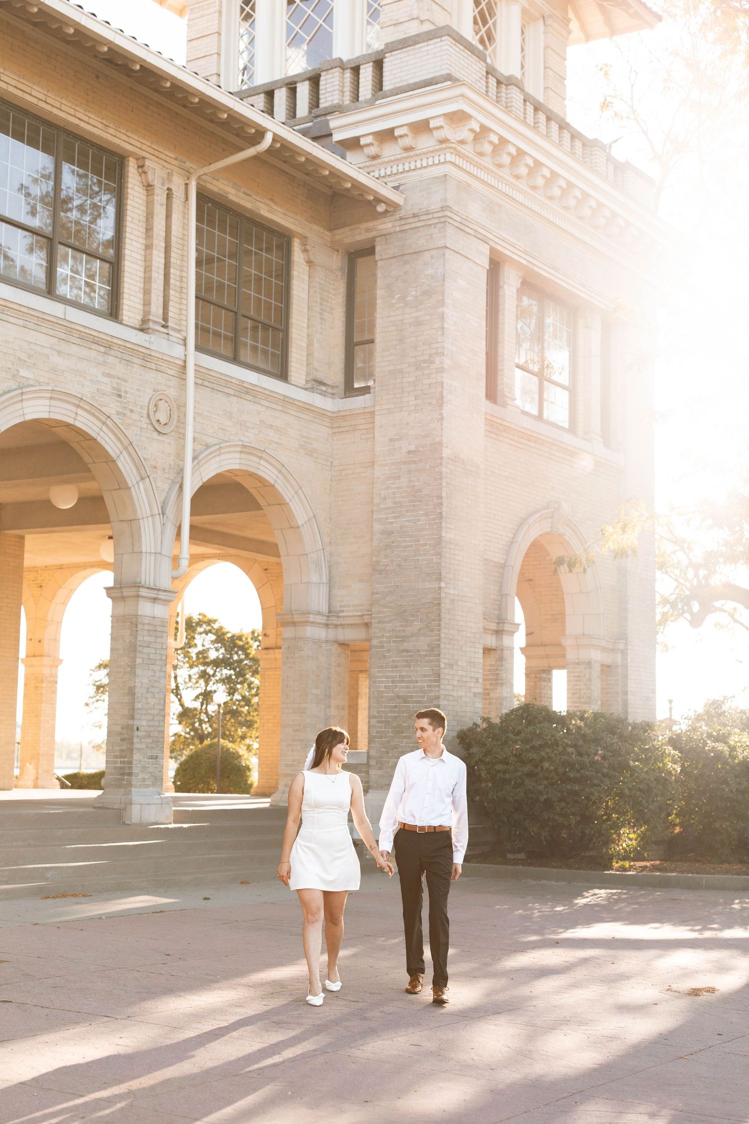 Couple holding hands walking in front of a sunlit historic stone building with arched windows.