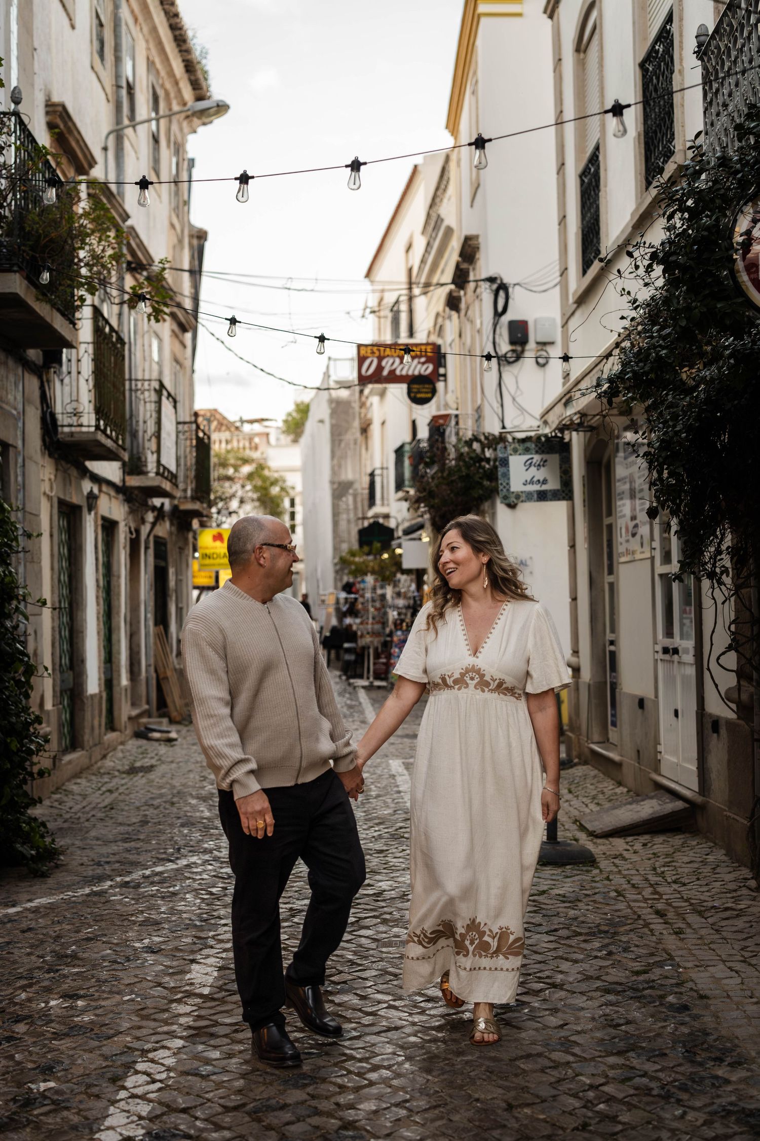 A couple walks down a charming cobblestone street in a historic European town with white buildings and string lights.