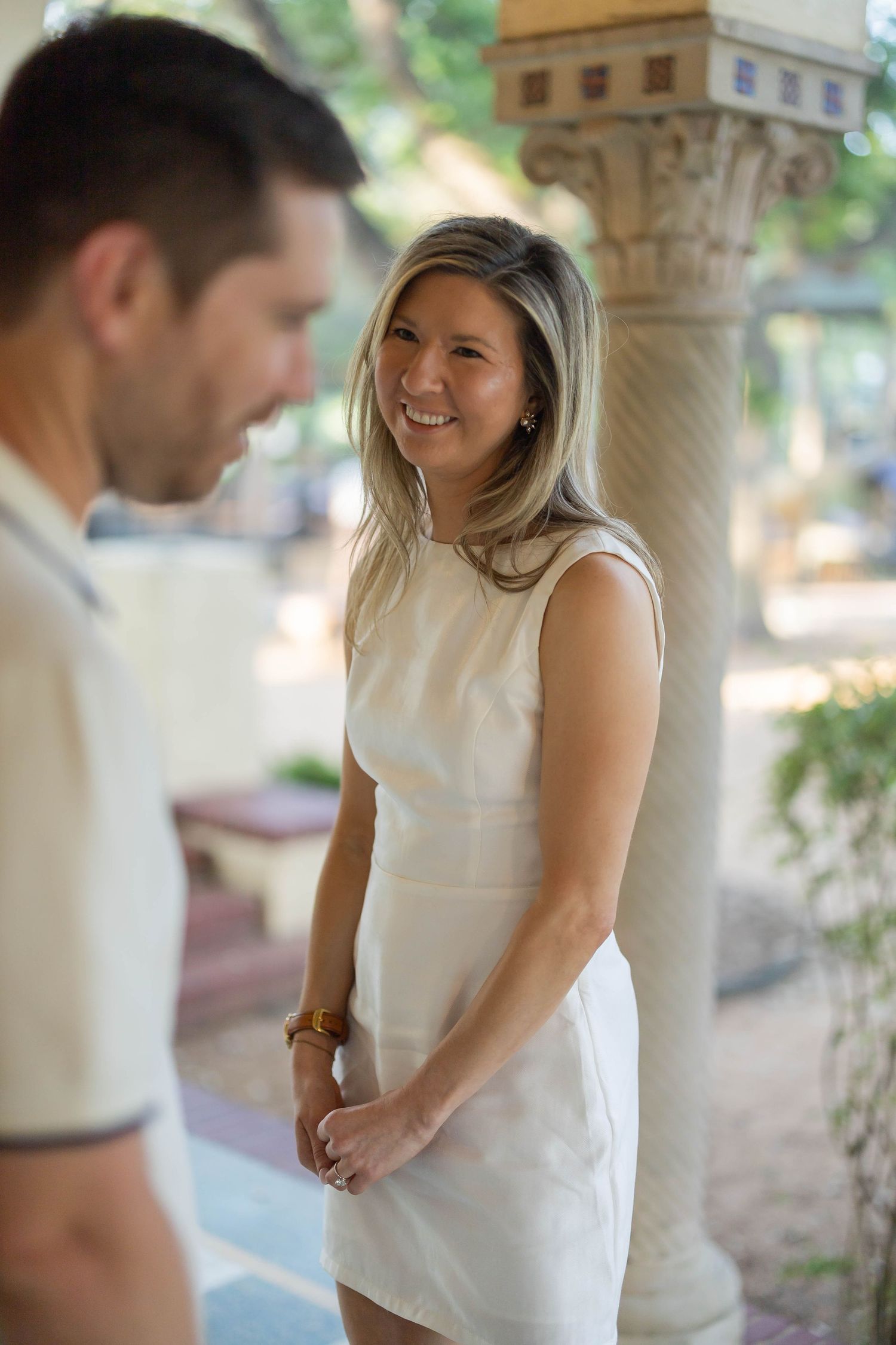 Smiling blonde woman in white dress standing by ornate column during outdoor engagement session.