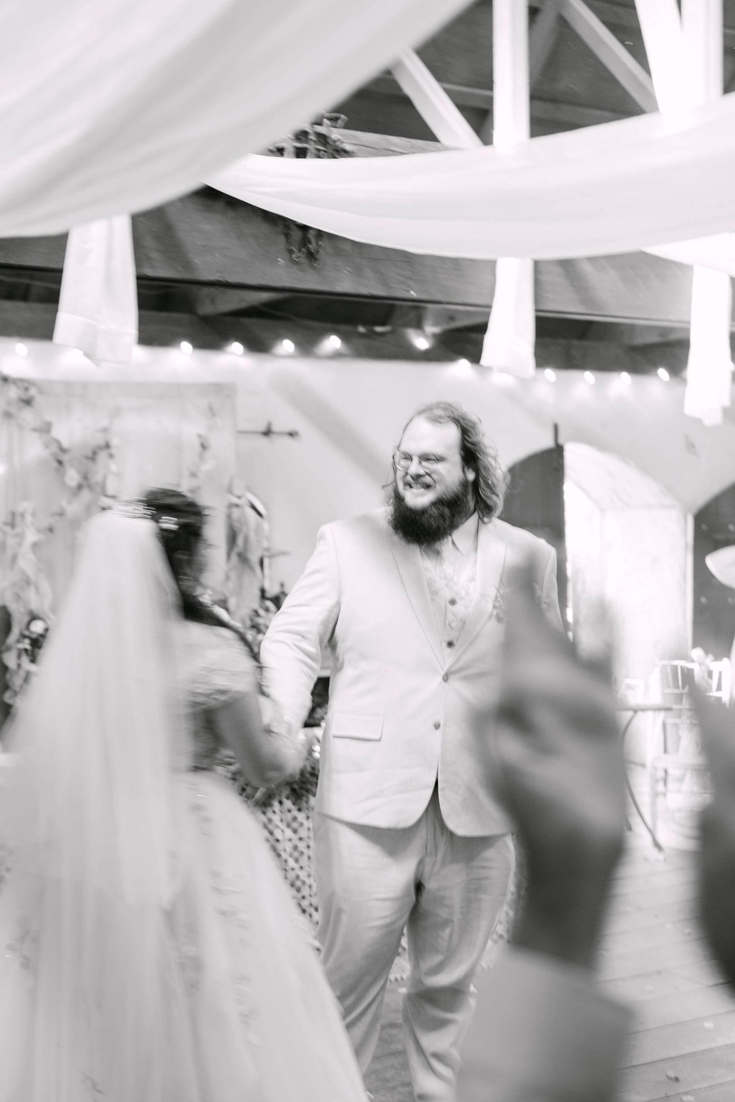 Black and white photo of a bearded groom in a light suit smiling at his bride during their first dance at wedding.