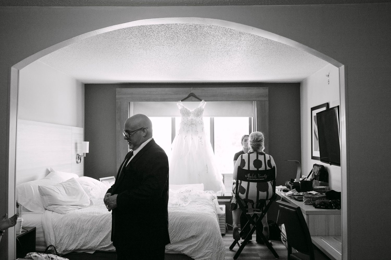 Black and white photo of a wedding dress hanging in a hotel room as a man stands nearby and a woman gets ready.