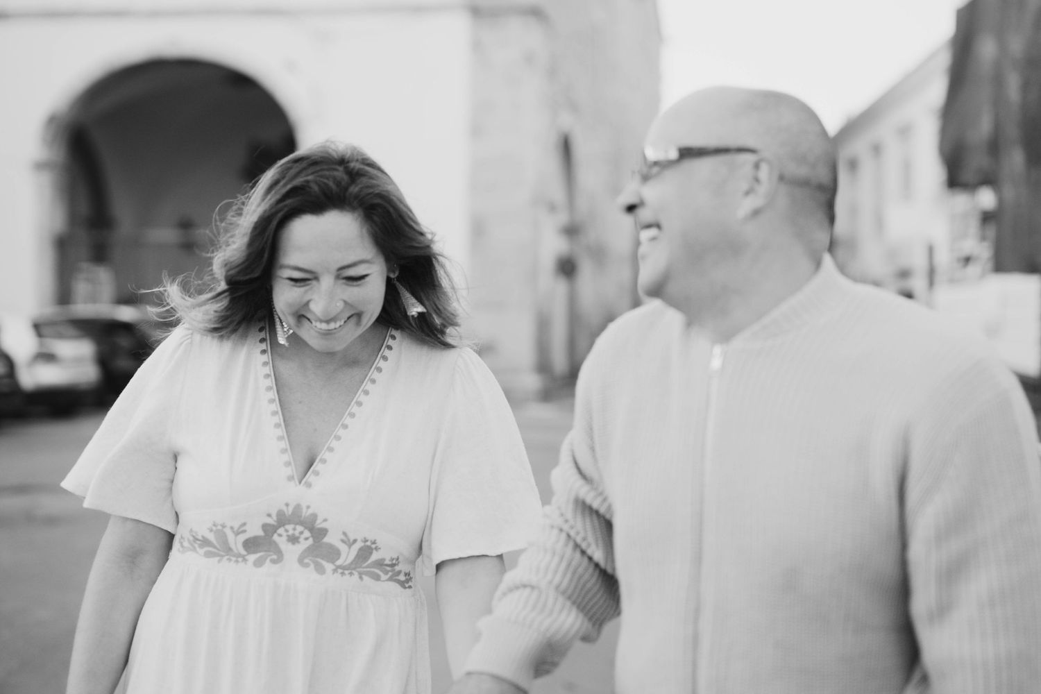 Black and white photo of a smiling couple holding hands and walking together outdoors near an archway.