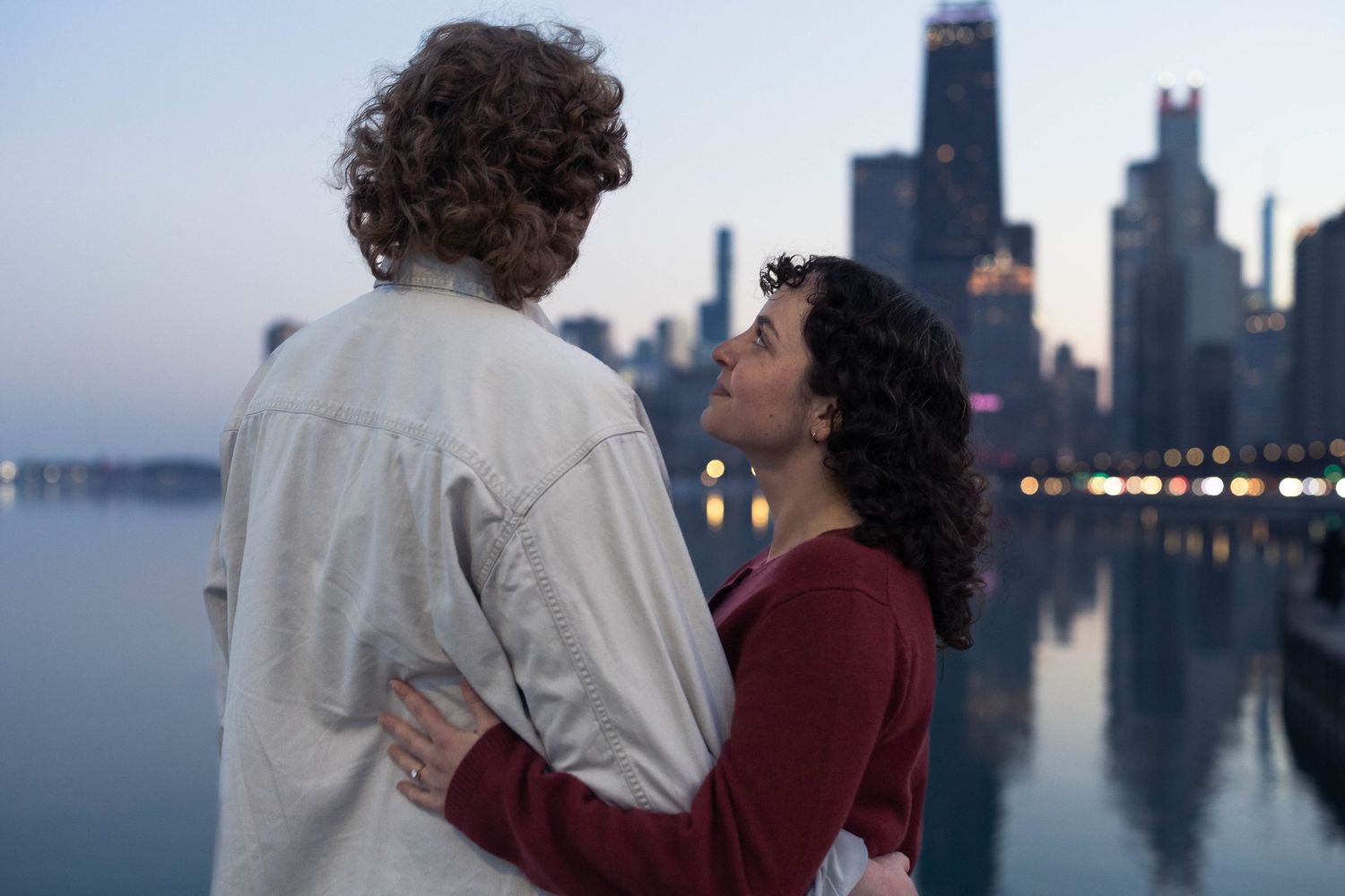 A couple embraces romantically by a city waterfront at dusk with a glowing skyline reflected in the water.